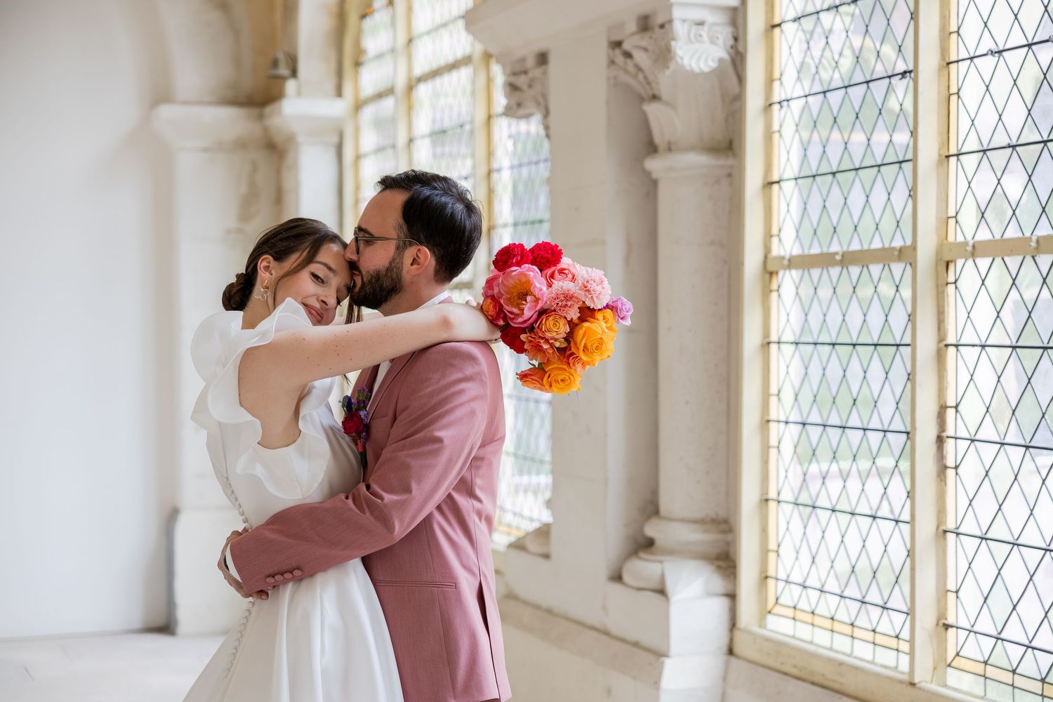 Bride and groom embracing tenderly in elegant hall with leaded windows, holding vibrant pink and orange bouquet.