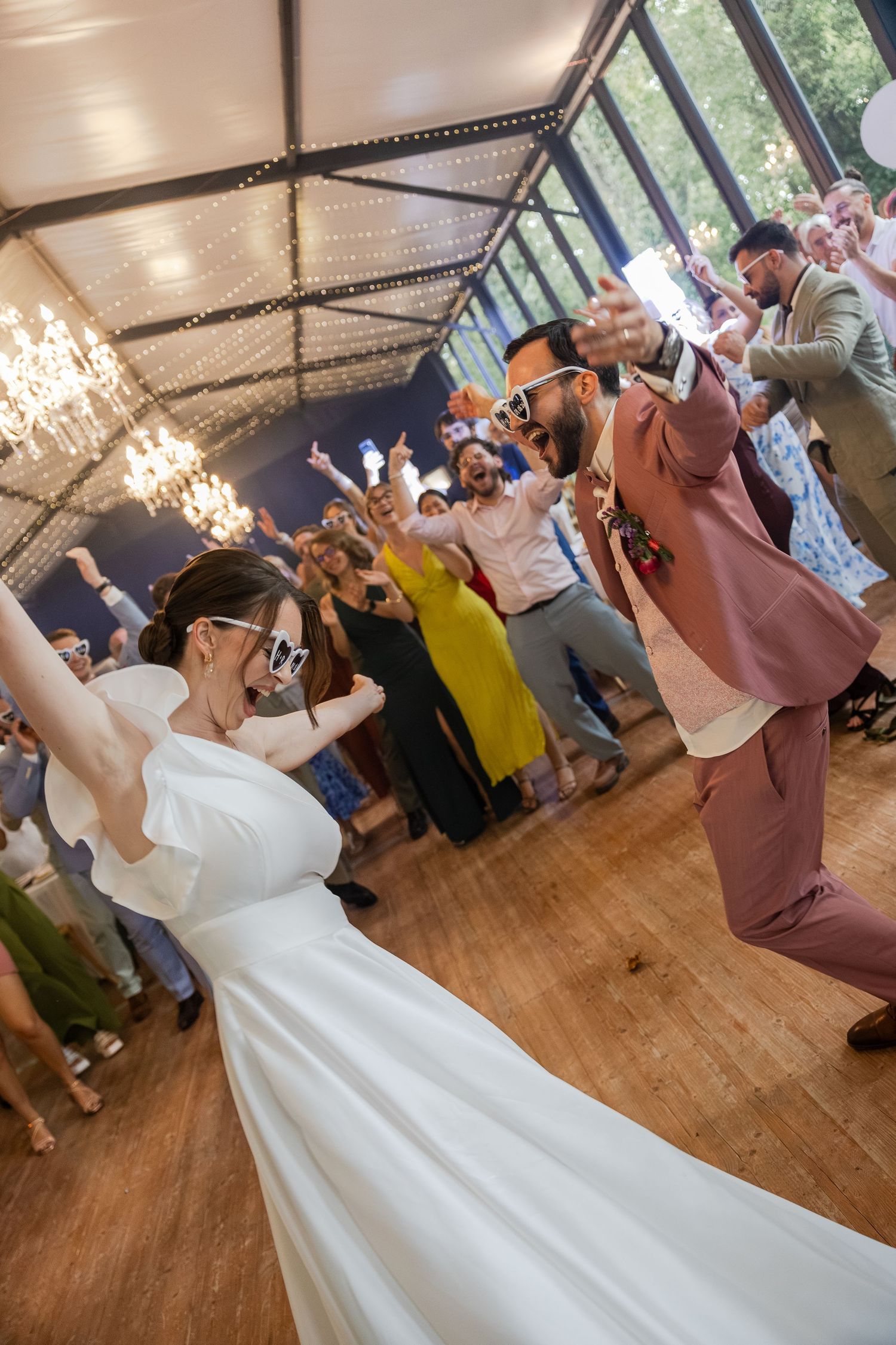 Bride and groom dancing joyfully in sunglasses during lively wedding reception on wooden dance floor.