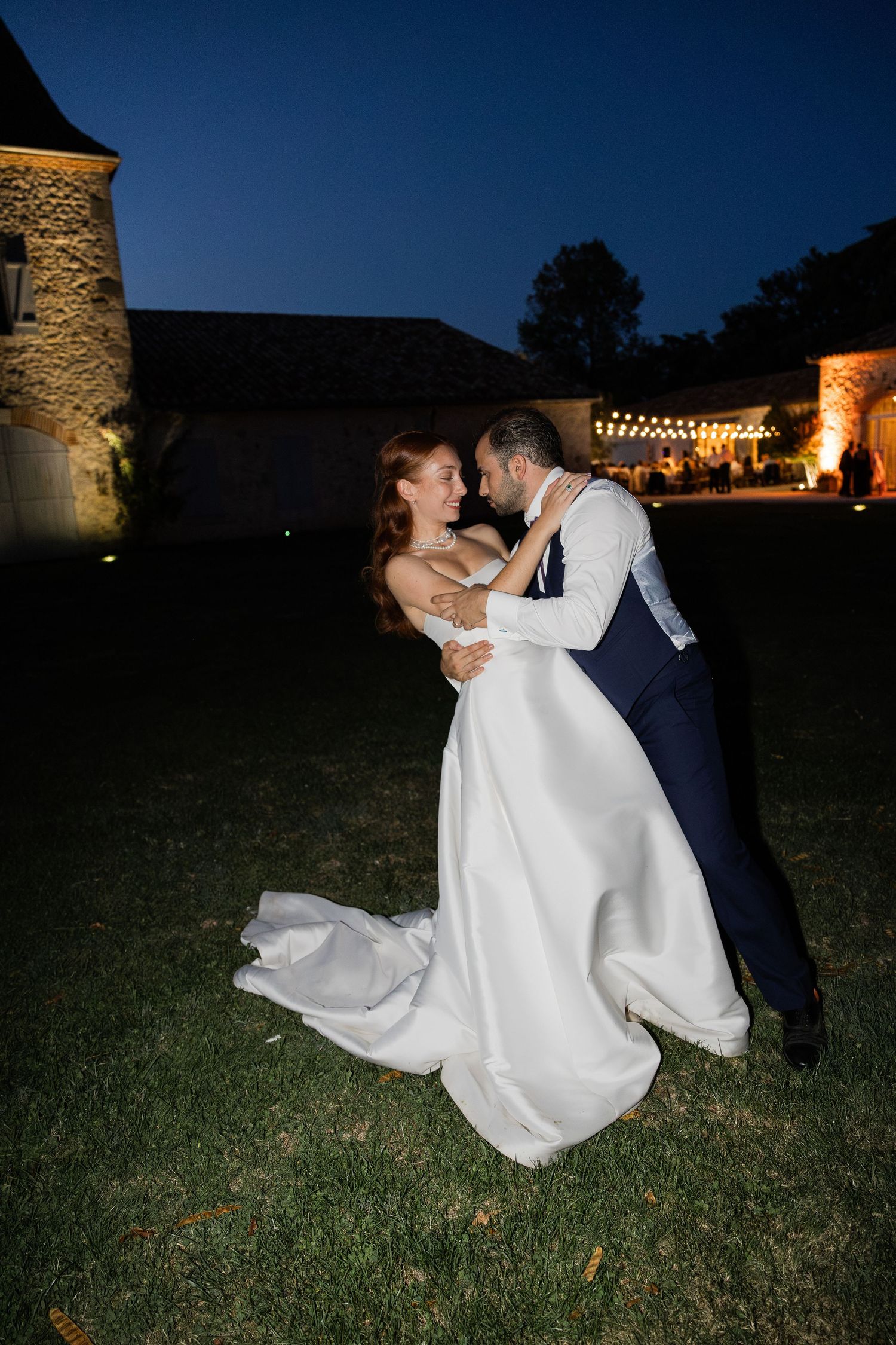 A bride in a white gown and groom in a navy suit share a romantic dance on a lawn at night.