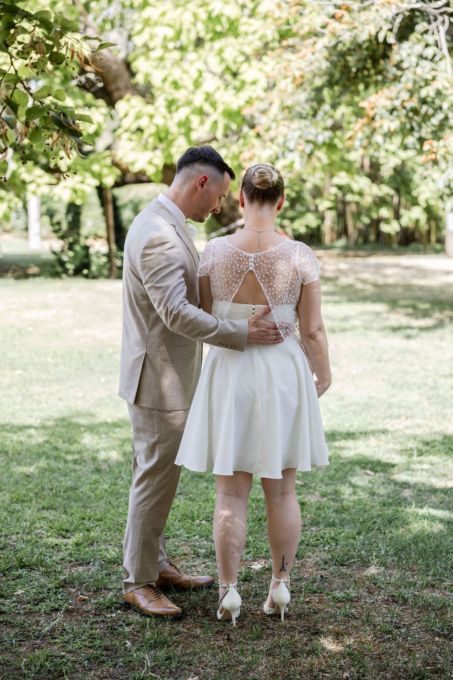 Couple in wedding attire standing in lush garden, groom in tan suit, bride in short white lace-back dress and heels.