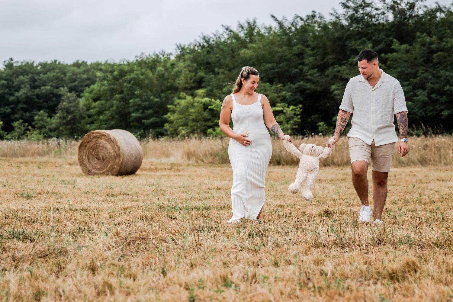 Pregnant woman and man walking in a field with hay bales, swinging a toddler between them on a sunny day.