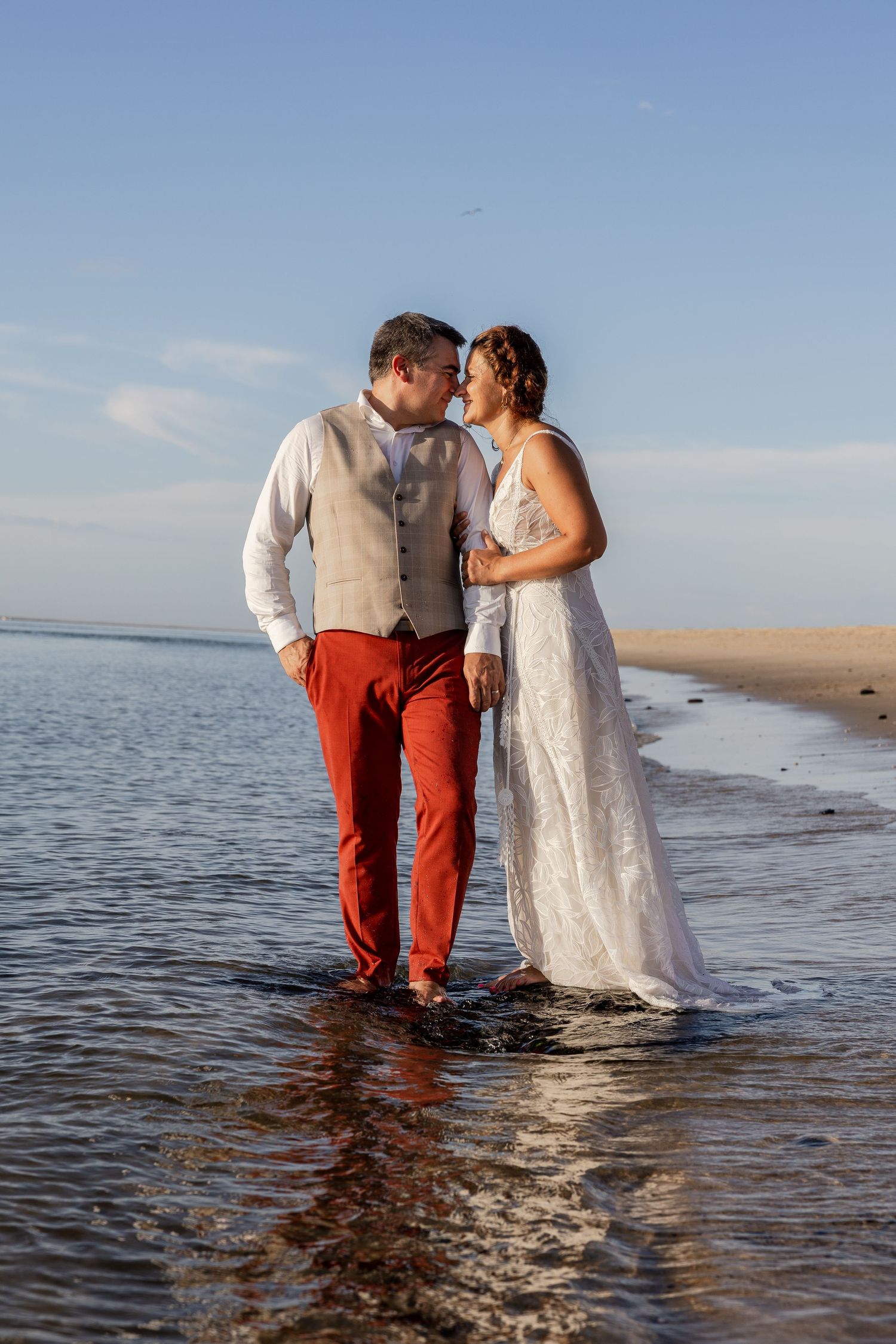 Couple in wedding attire standing barefoot in shallow ocean water, sharing a romantic moment at sunset on a sandy beach.