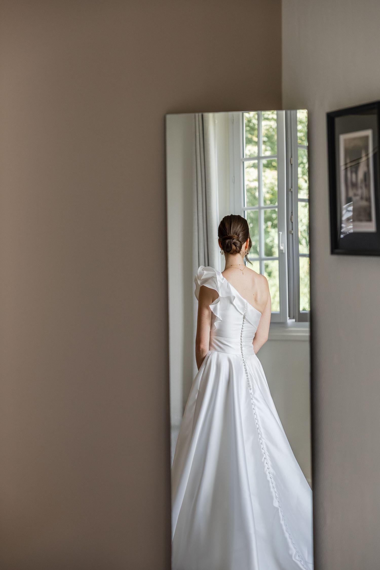 Bride in elegant one-shoulder white wedding gown with ruffle detail, viewed from behind near a floor-length mirror.