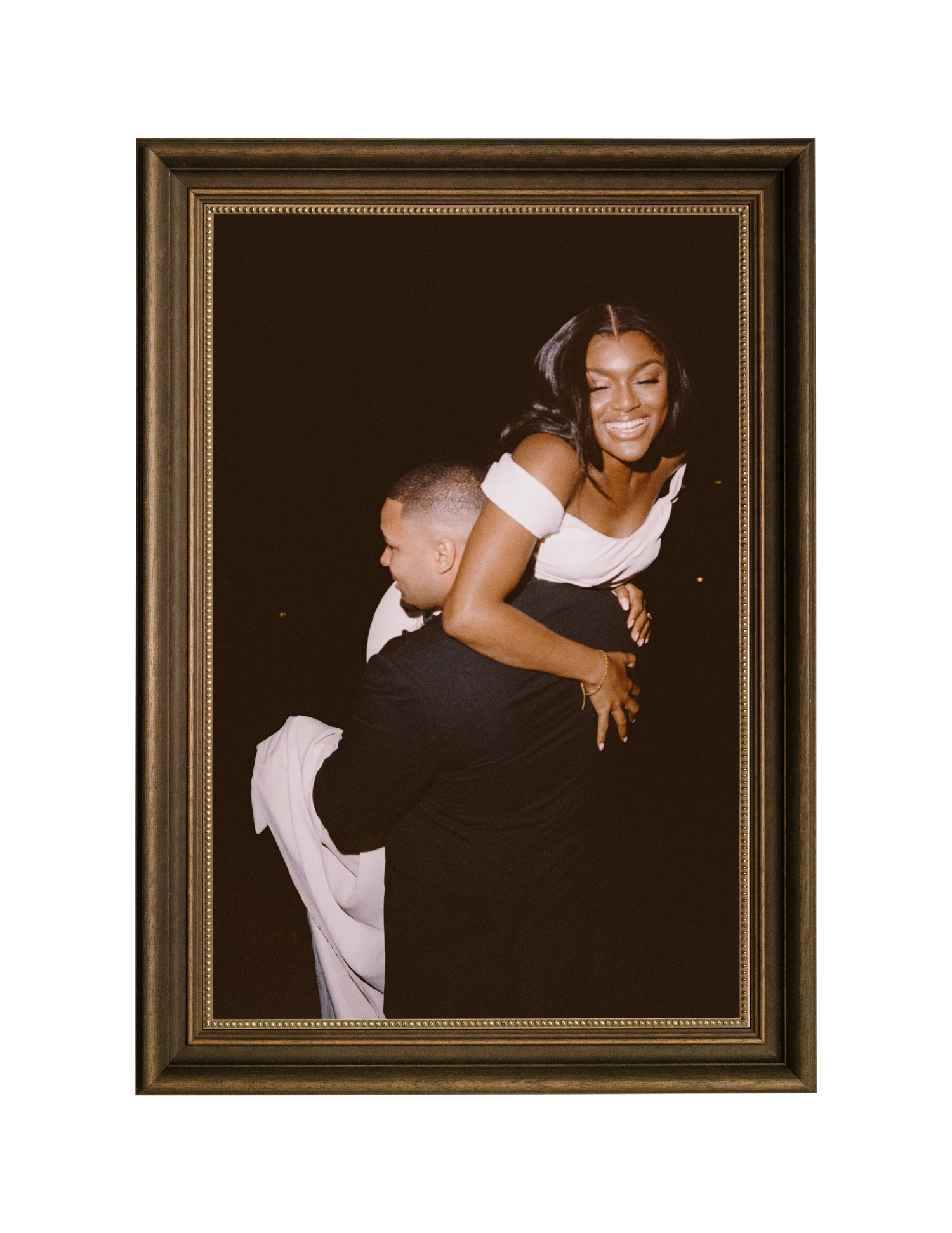A joyful couple embraces in a framed portrait, woman in white dress laughing as man lifts her against dark background.