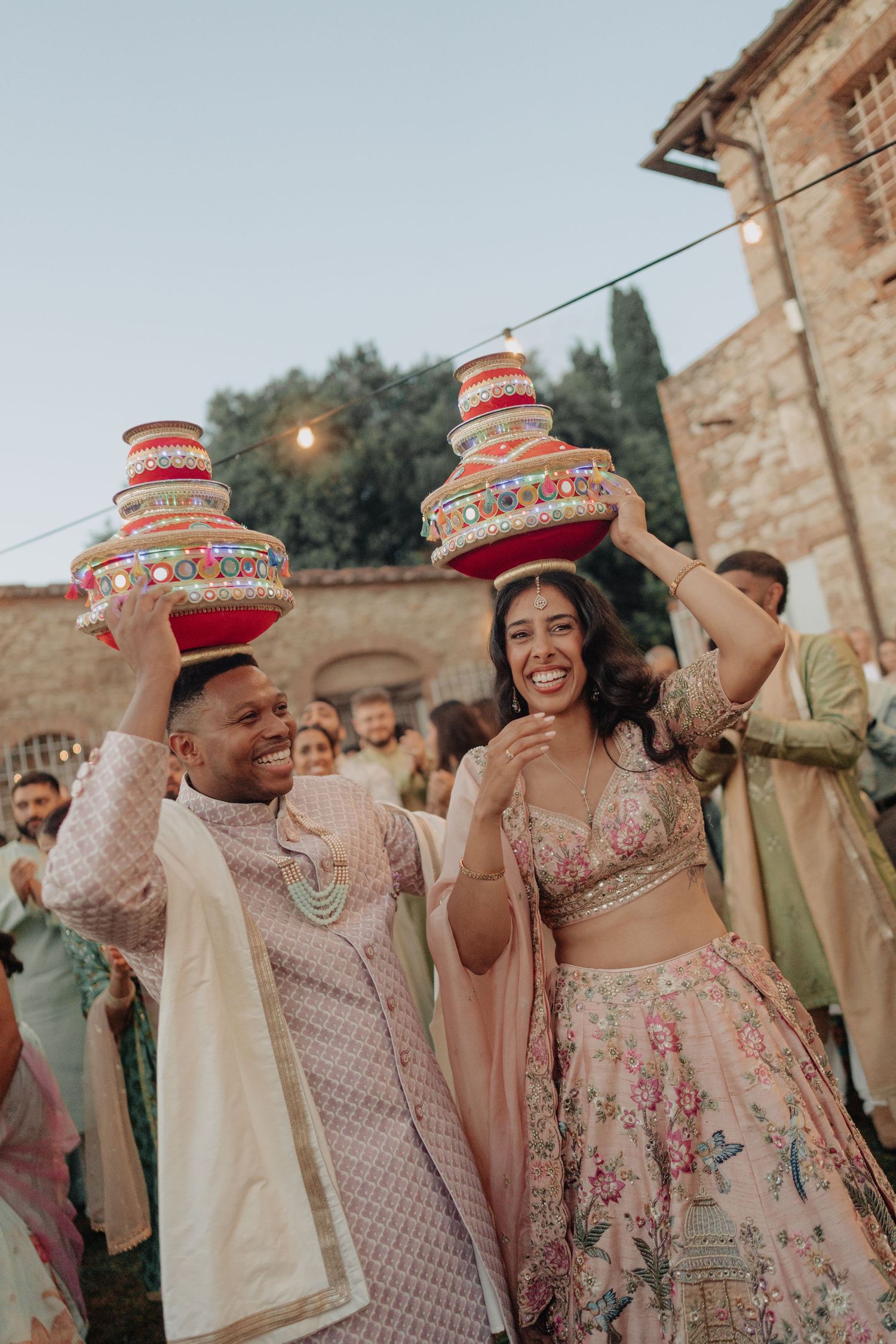 Couple in Indian wedding attire balancing decorated pots on their heads during a colorful outdoor ceremony.