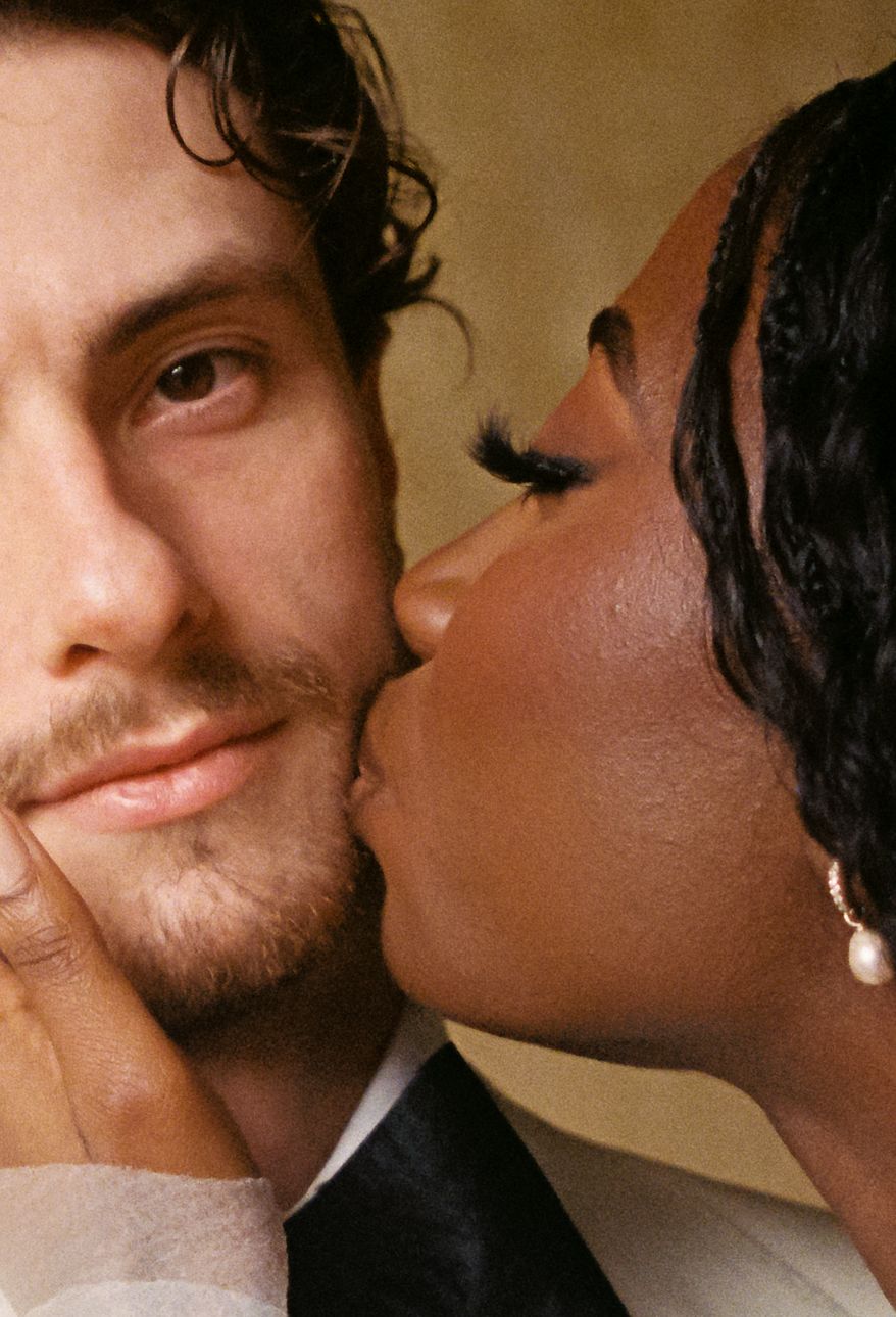 A woman kisses the cheek of a man in a close romantic portrait, both dressed elegantly.