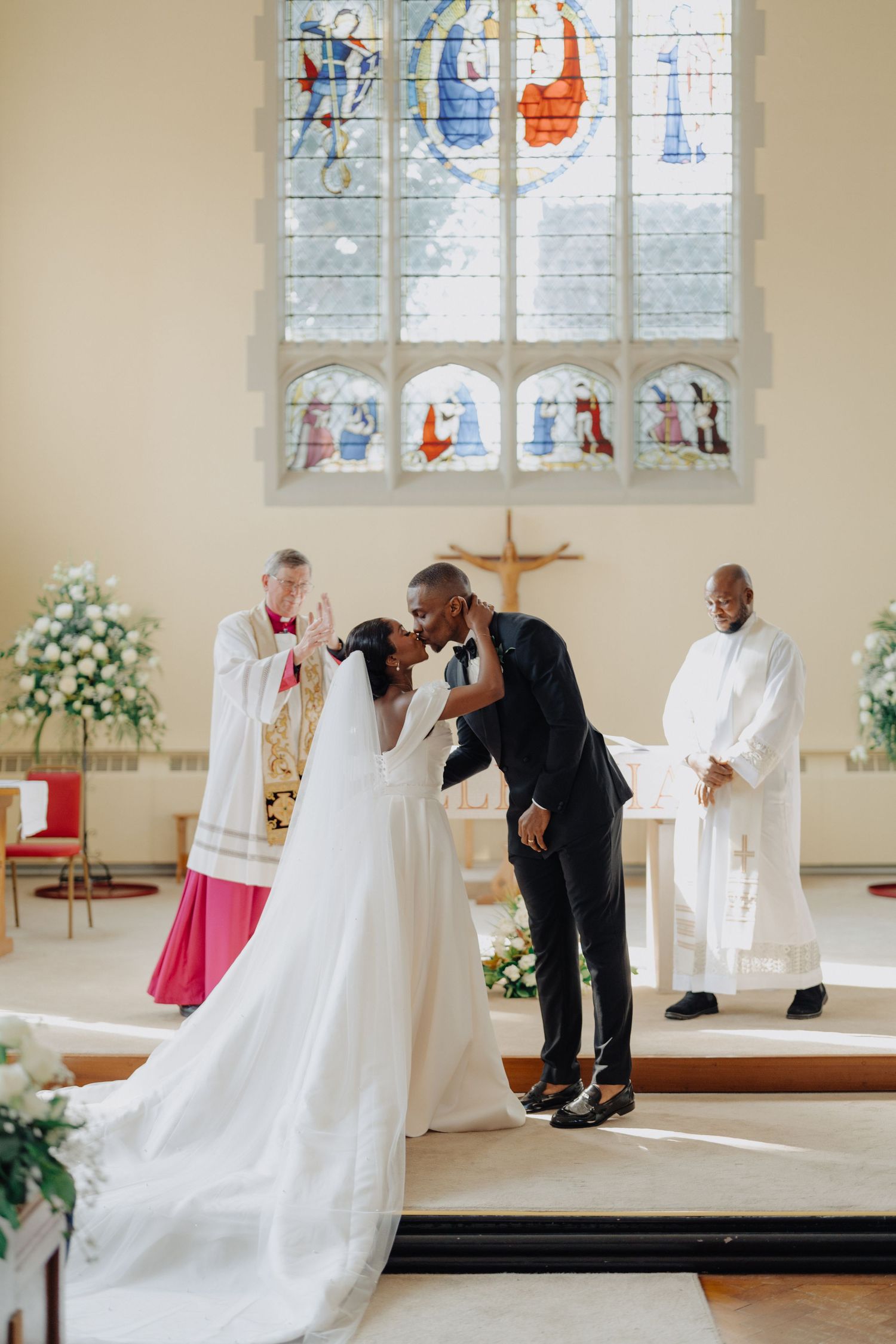Bride and groom share first kiss at church altar during wedding ceremony with stained glass windows and officiant behind them.