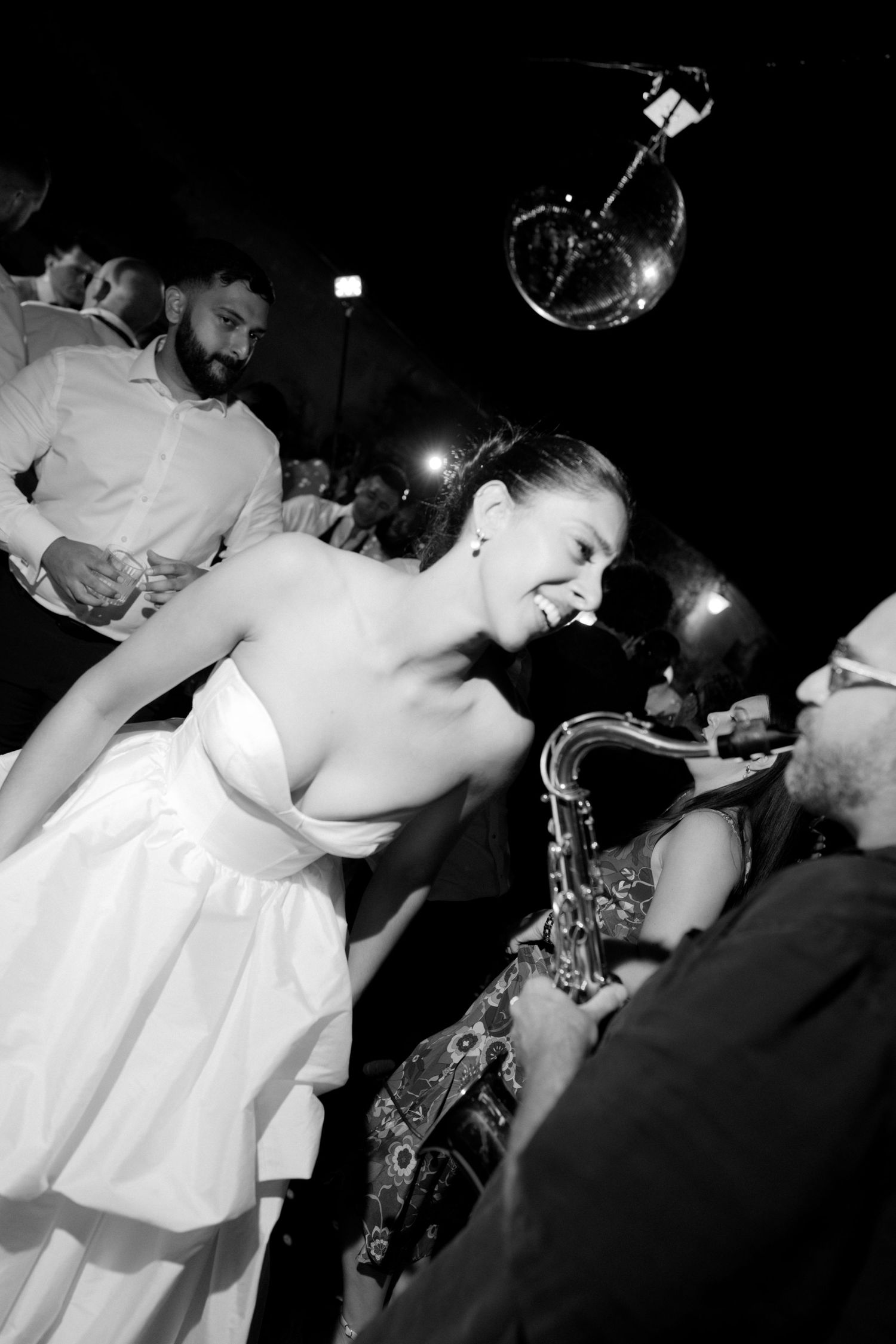 Bride in white dress dances with saxophone player at wedding reception under disco ball, black and white photo.