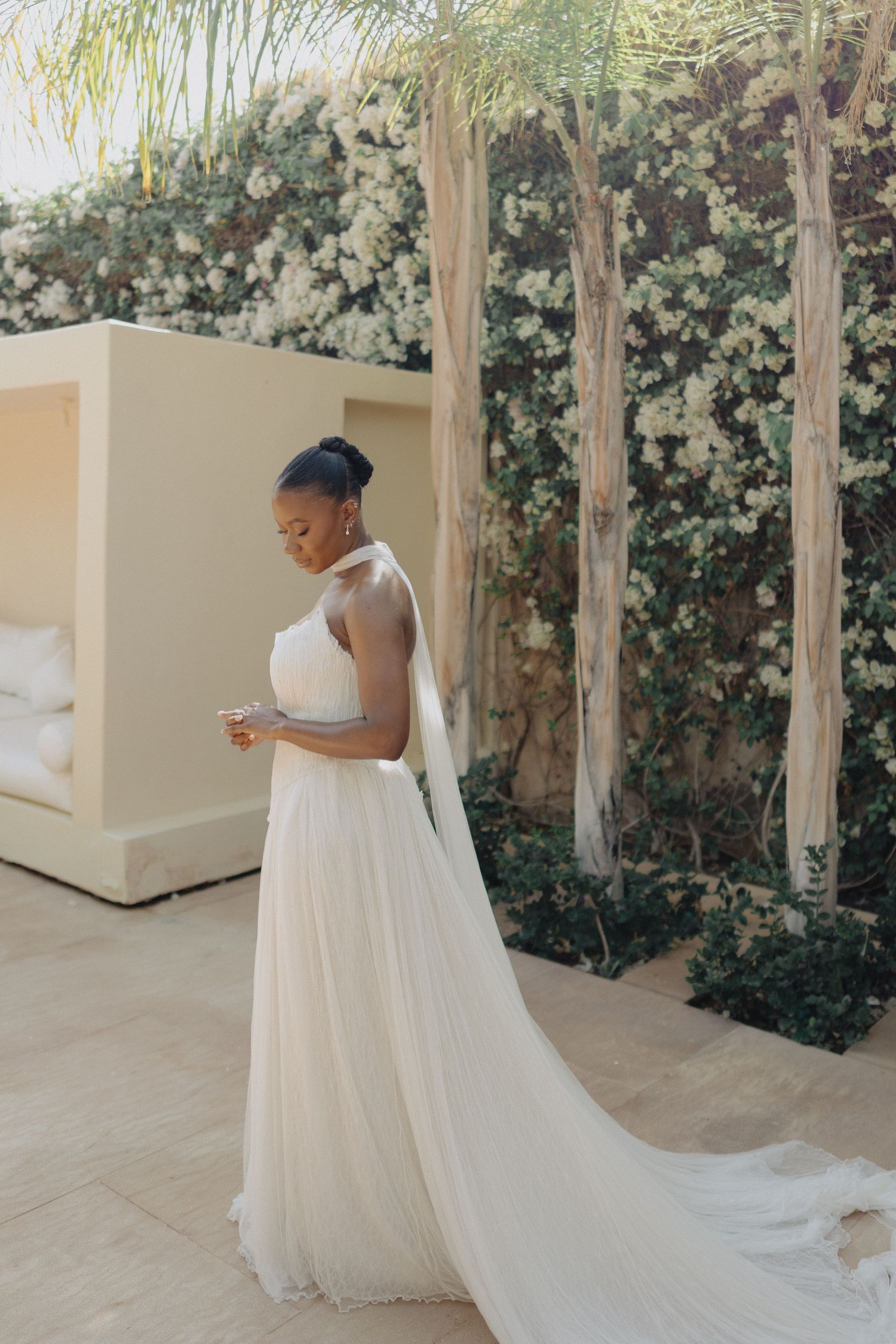 Elegant bride in a white halter gown with flowing train stands in a sunlit garden with blooming white flowers.