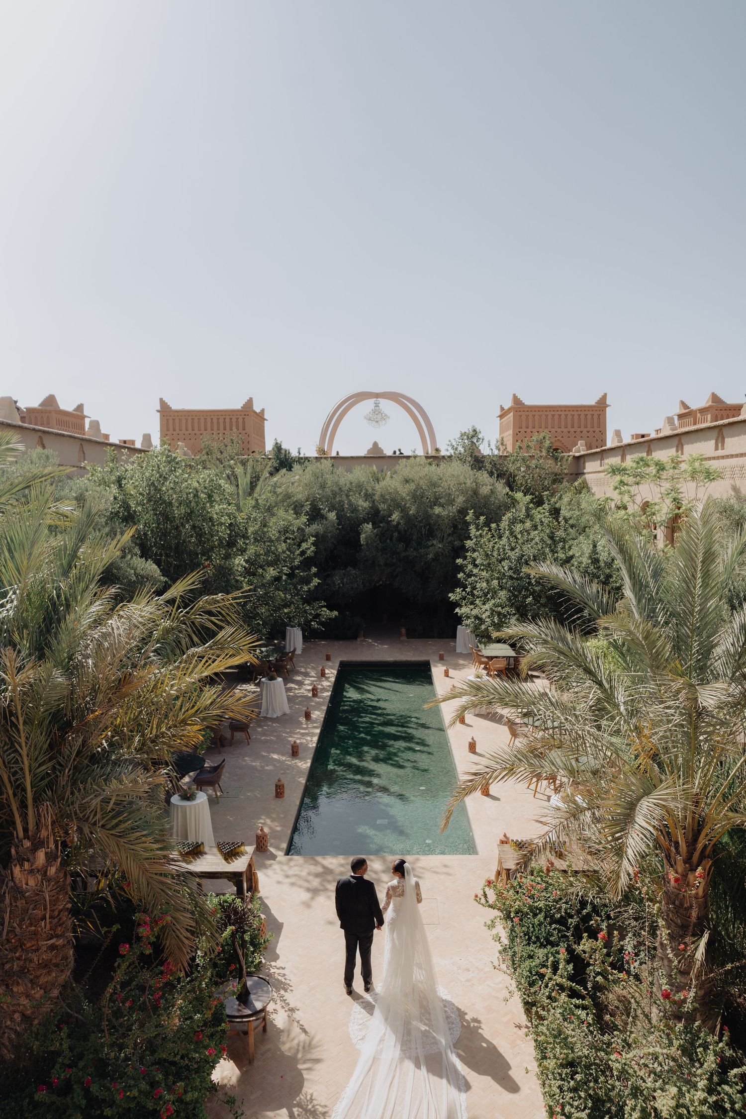 Bride and groom walk toward a pool surrounded by palm trees at a Moroccan wedding venue under a clear sky.