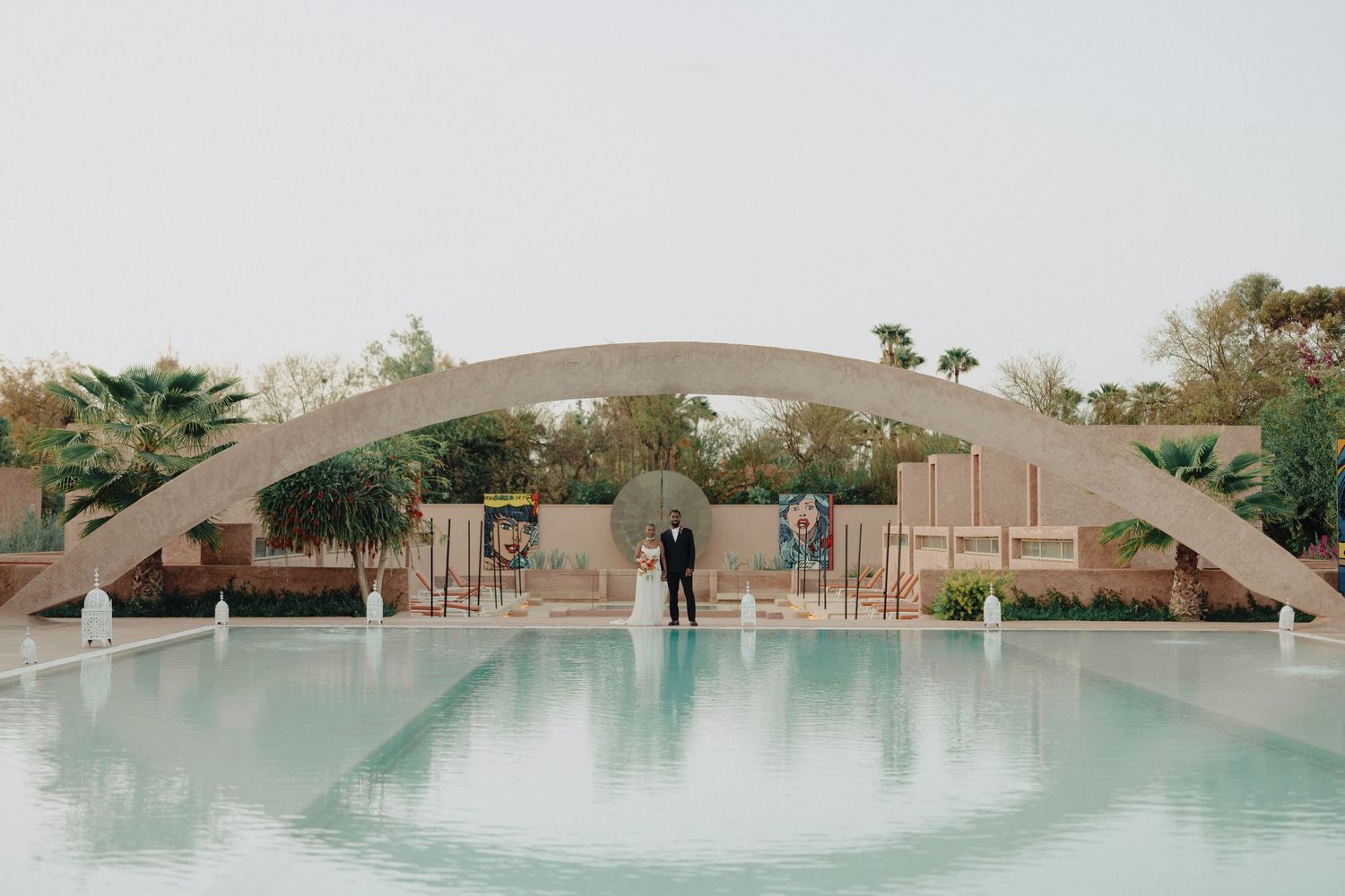 Couple standing by a serene pool beneath a grand arch at an elegant outdoor wedding venue.