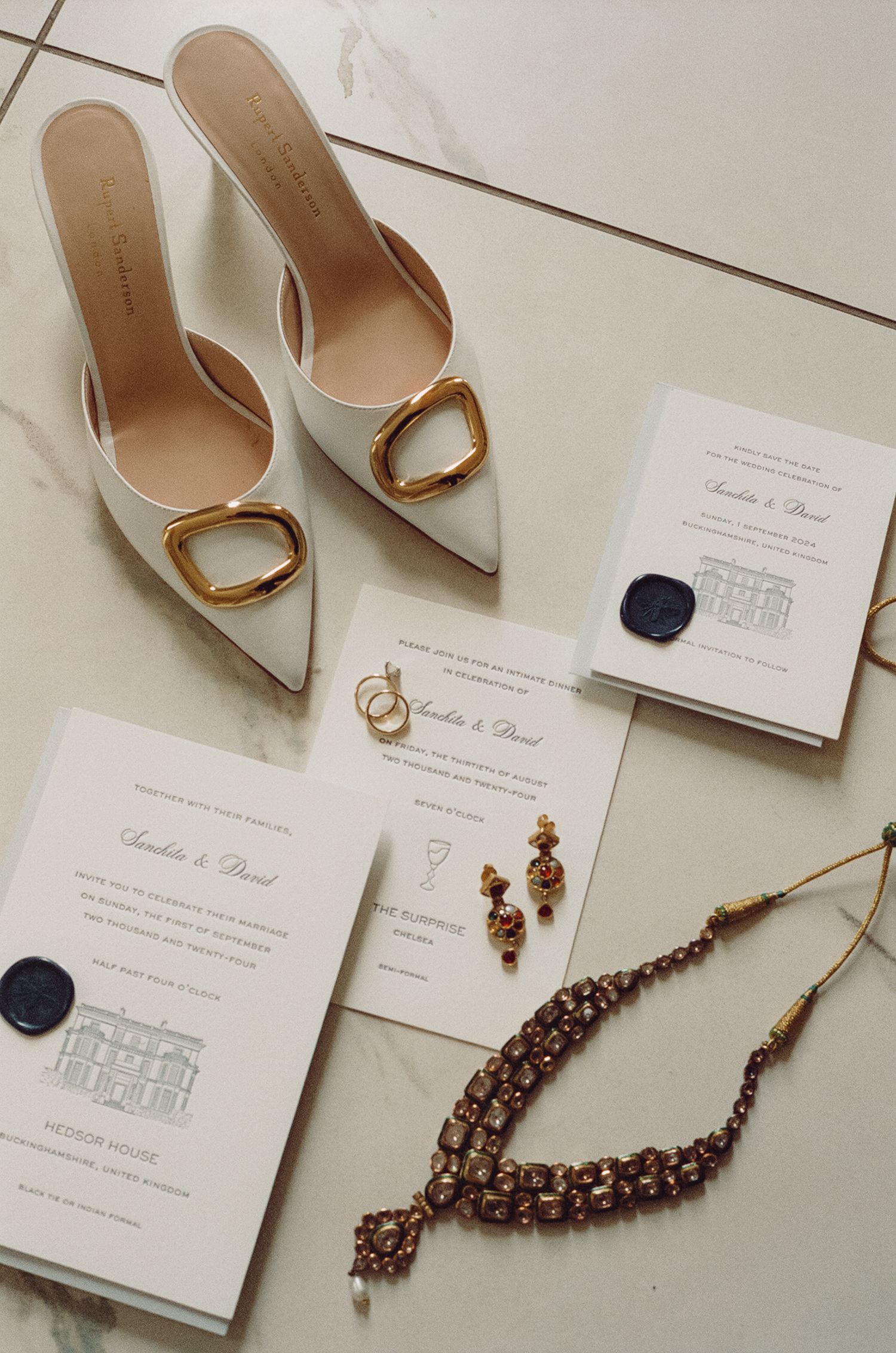Elegant wedding flat lay featuring white gold-buckle heels, floral invitations, gold rings, garnet drop earrings, and a beaded necklace.