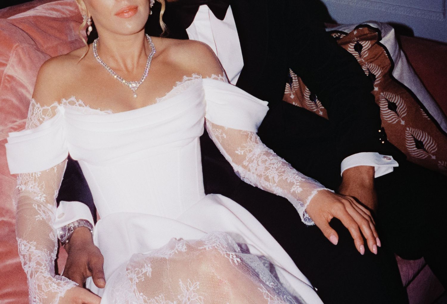 Bride in white off-shoulder lace wedding gown with elbow gloves and diamond necklace seated beside groom in tuxedo.