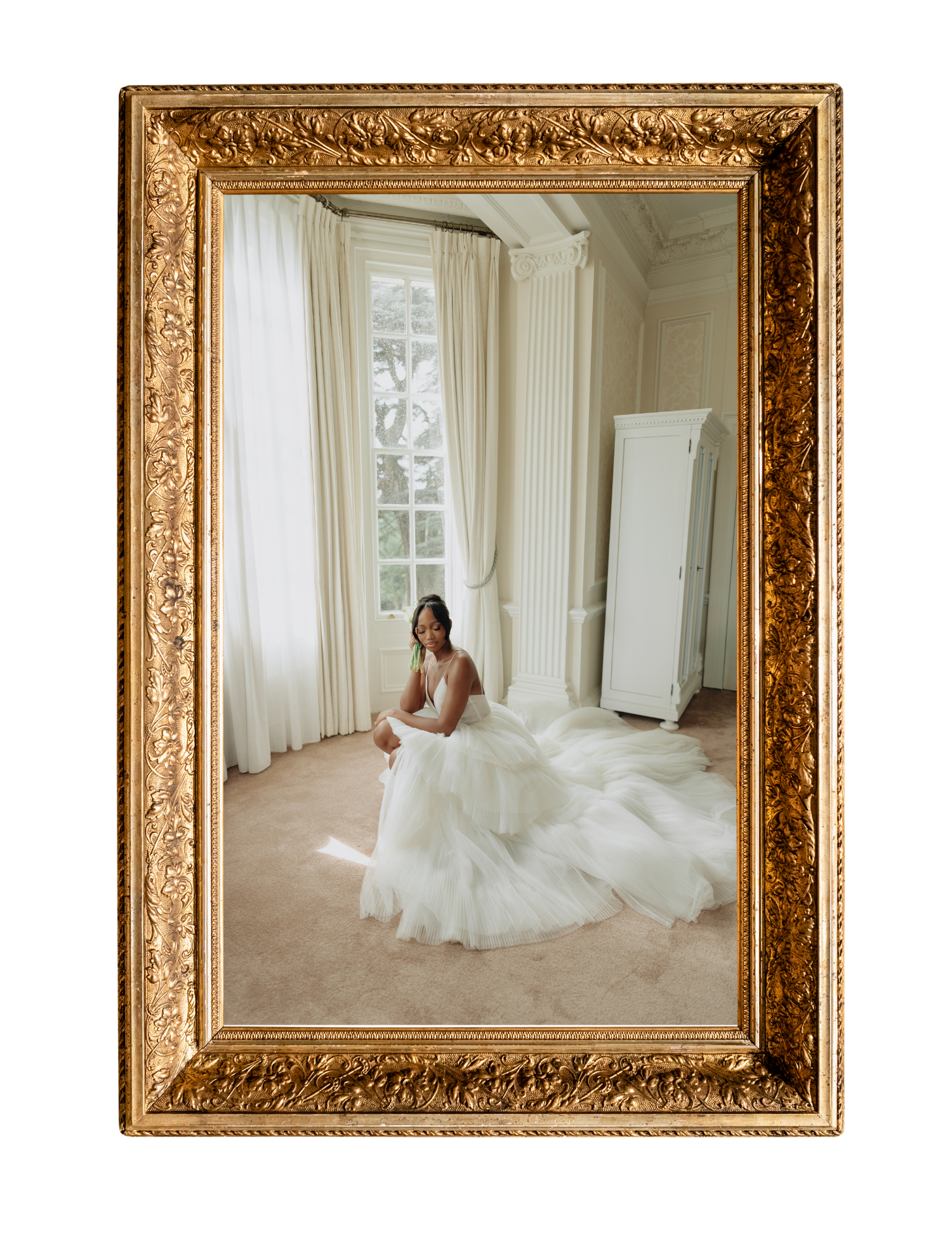 Bride in a flowing white tulle gown seated gracefully in a sunlit room, framed by an ornate gold mirror.