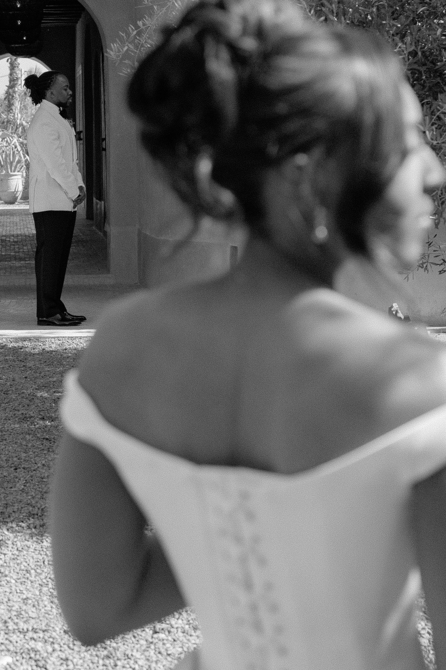 Black and white wedding photo of bride in off-shoulder gown, groom standing in doorway in background.
