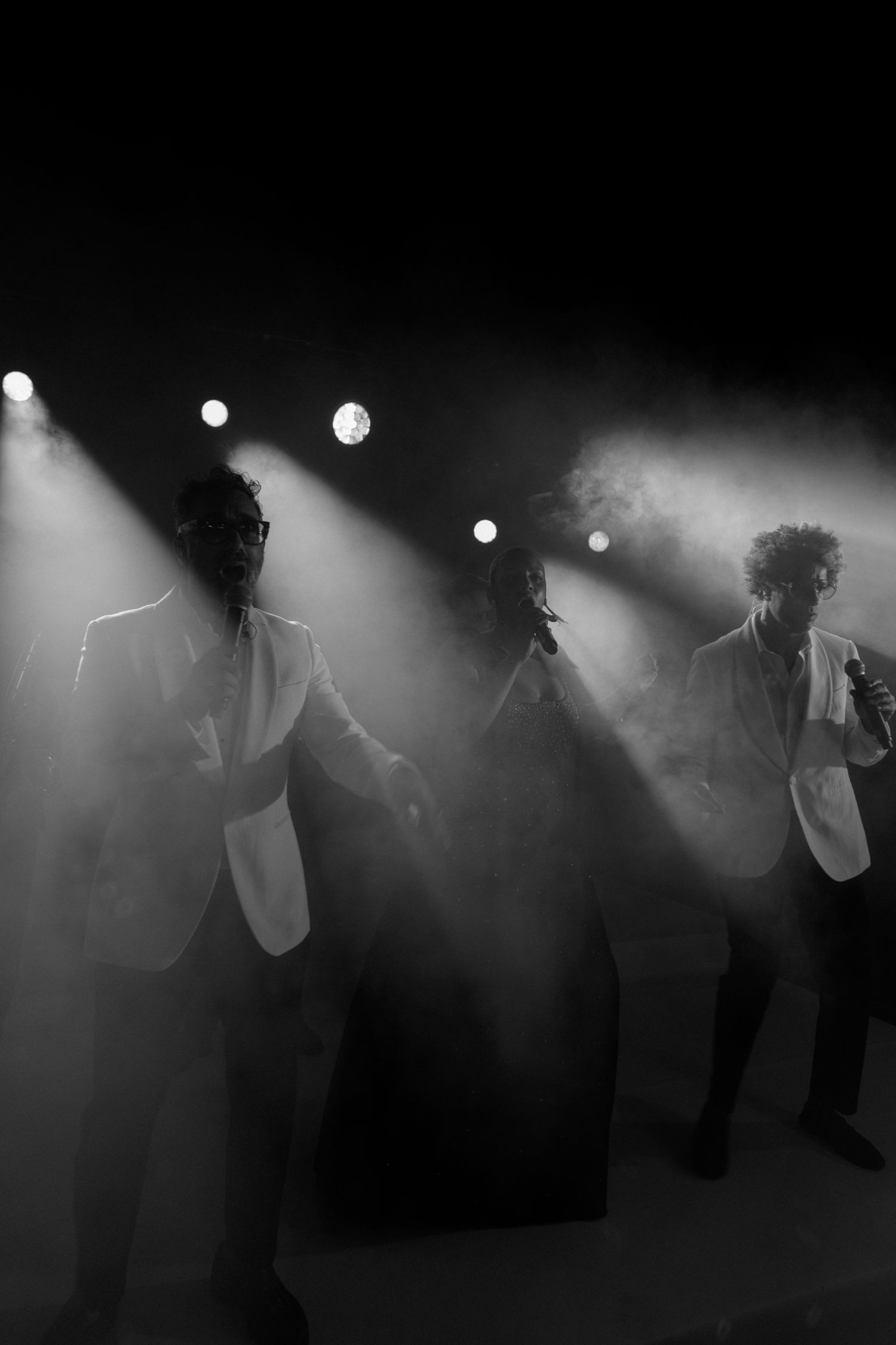 Three performers in white suits dance on a smoky stage under dramatic spotlights in a black and white concert photo.