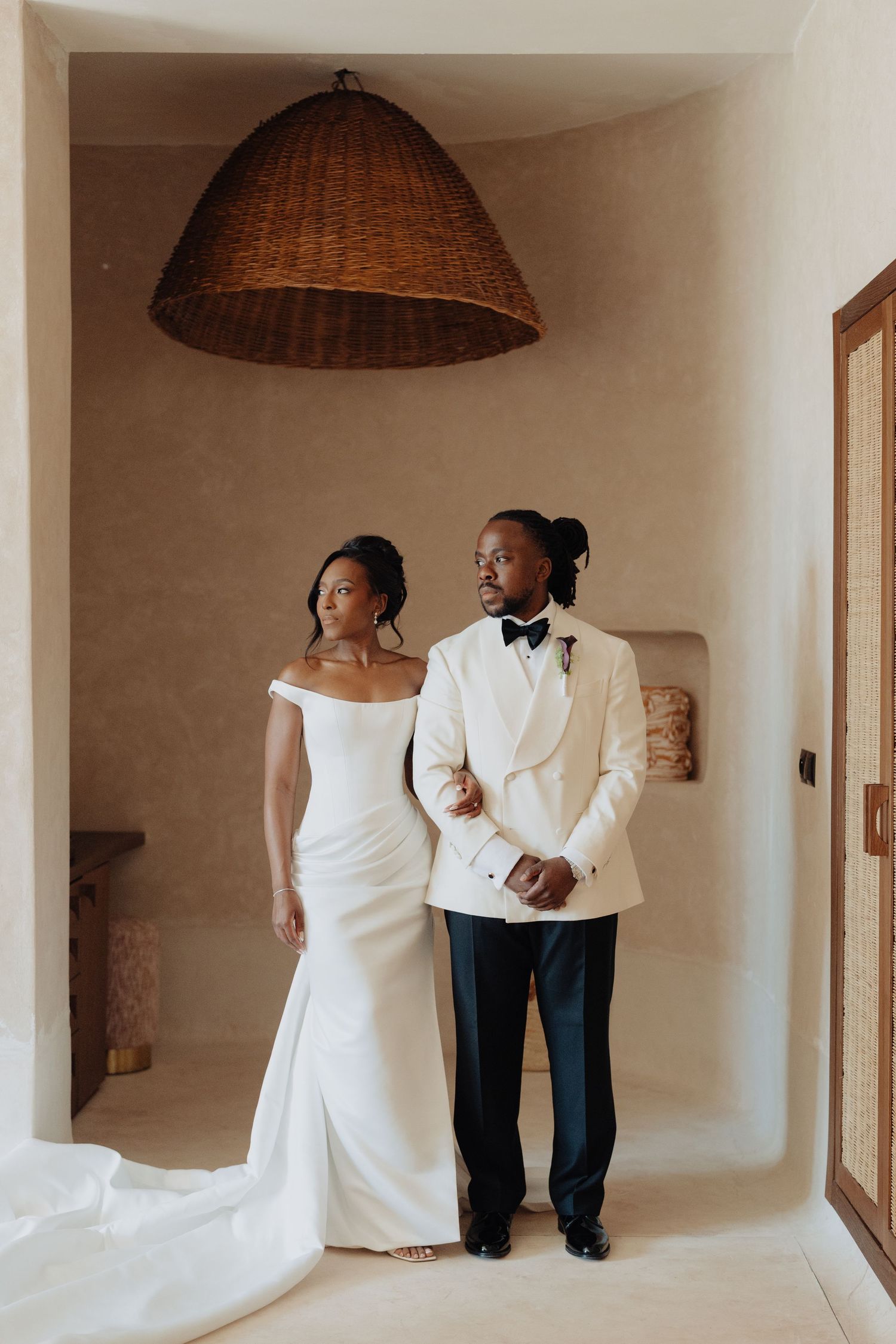 Elegant couple in wedding attire, bride in off-shoulder gown and groom in white tuxedo, posing under woven pendant light.