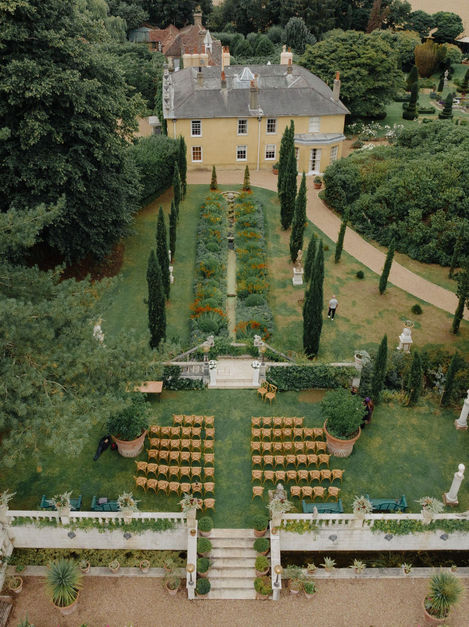 Aerial view of an elegant English manor house wedding ceremony with golden chairs arranged on manicured formal gardens.