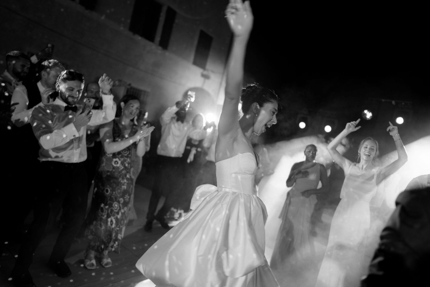 Black and white photo of a bride dancing joyfully with guests celebrating at an outdoor wedding reception at night.