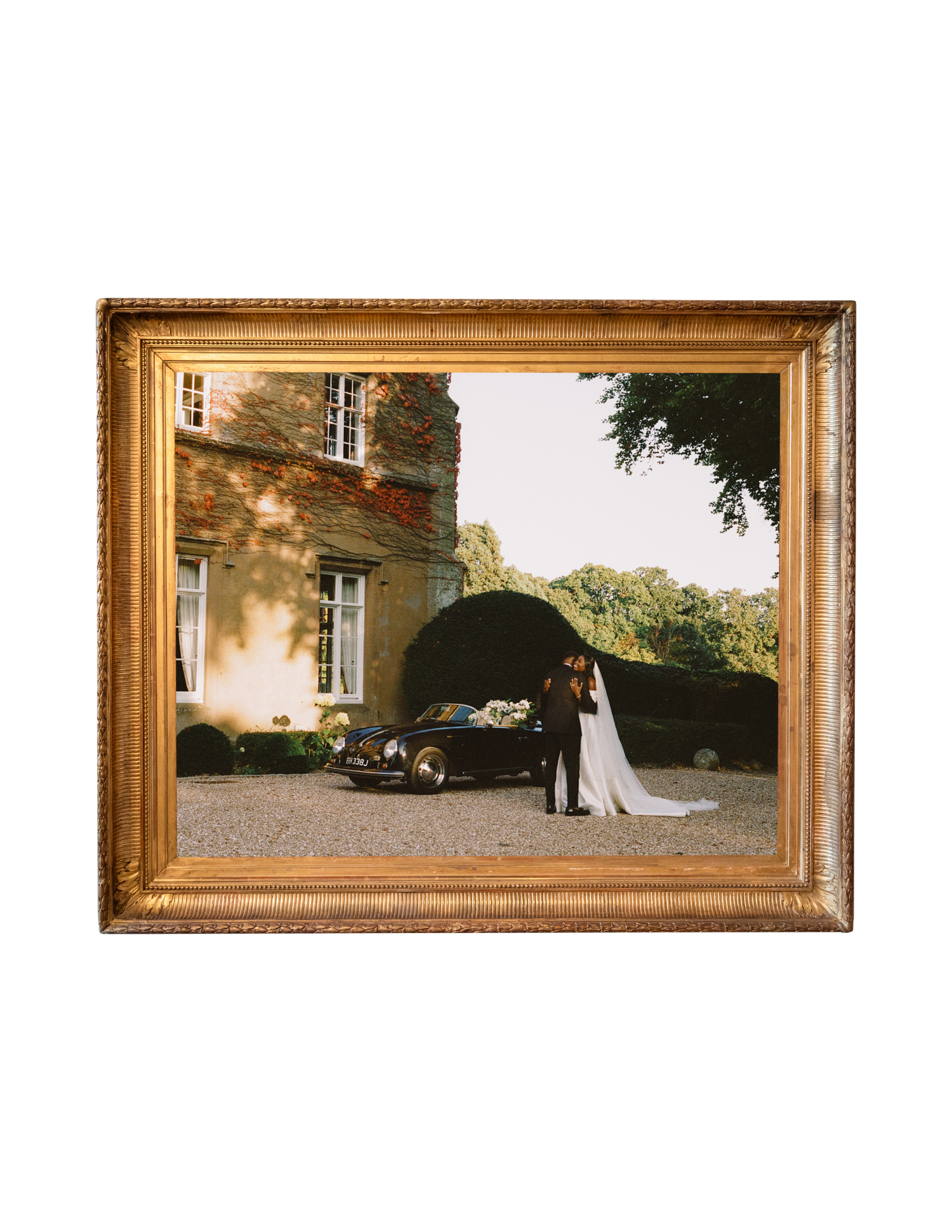 Gold-framed wedding photo of bride and groom beside vintage car outside elegant French château.
