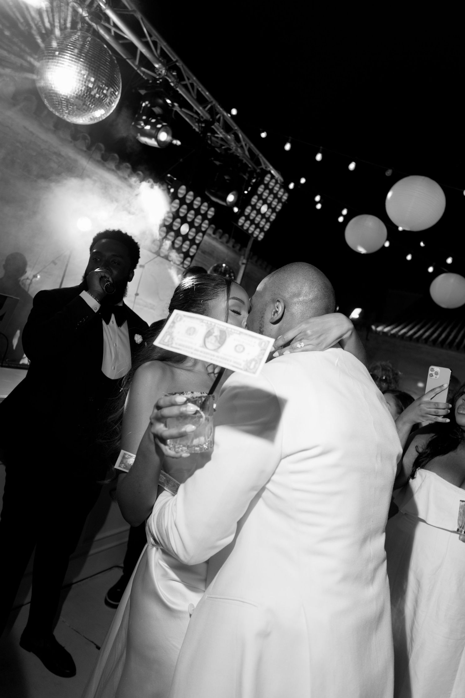 Black and white wedding dance photo with couple kissing, dollar bill, disco ball, and balloons at elegant reception.