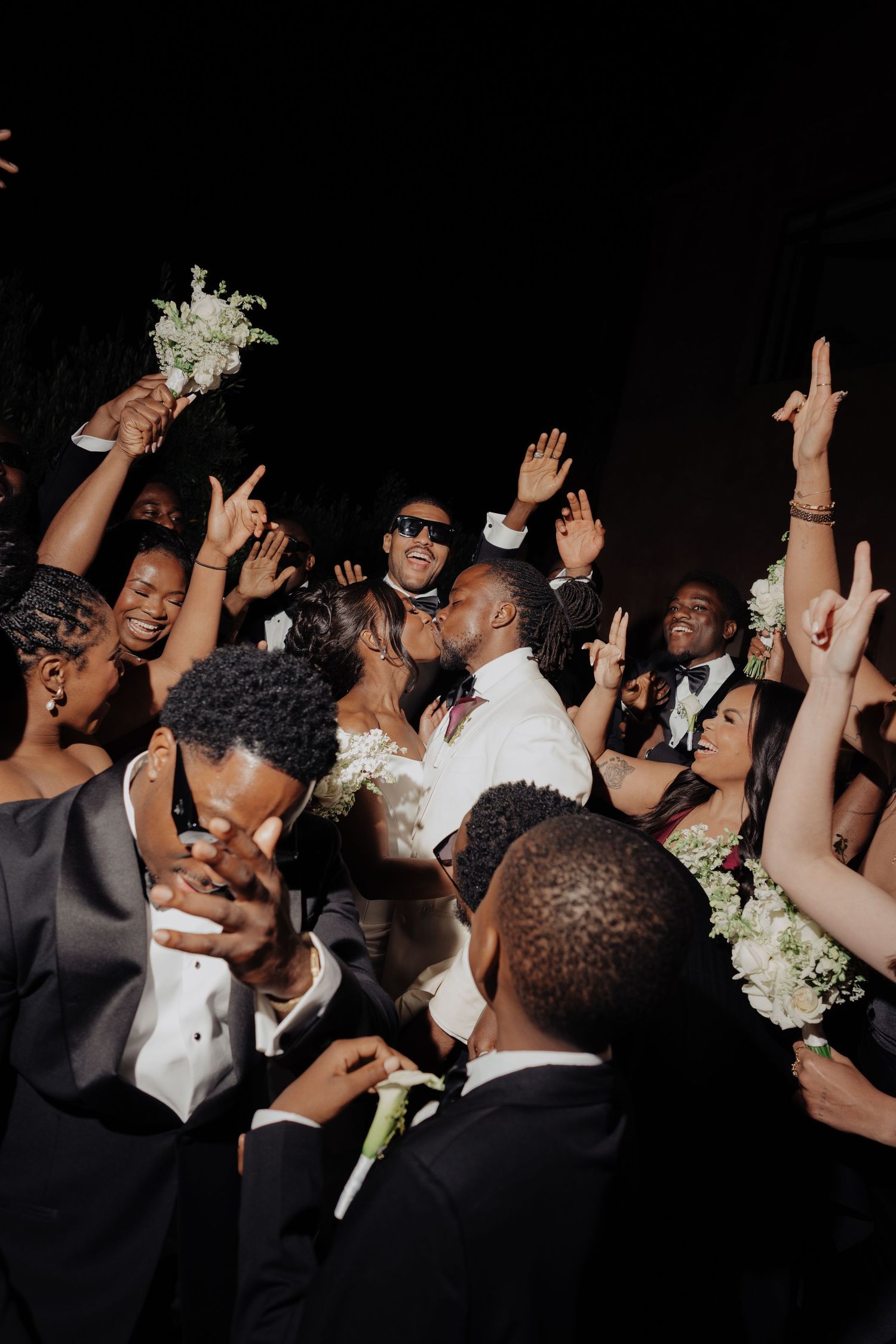Joyful wedding party celebration with bride and groom kissing surrounded by guests raising hands and bouquets.