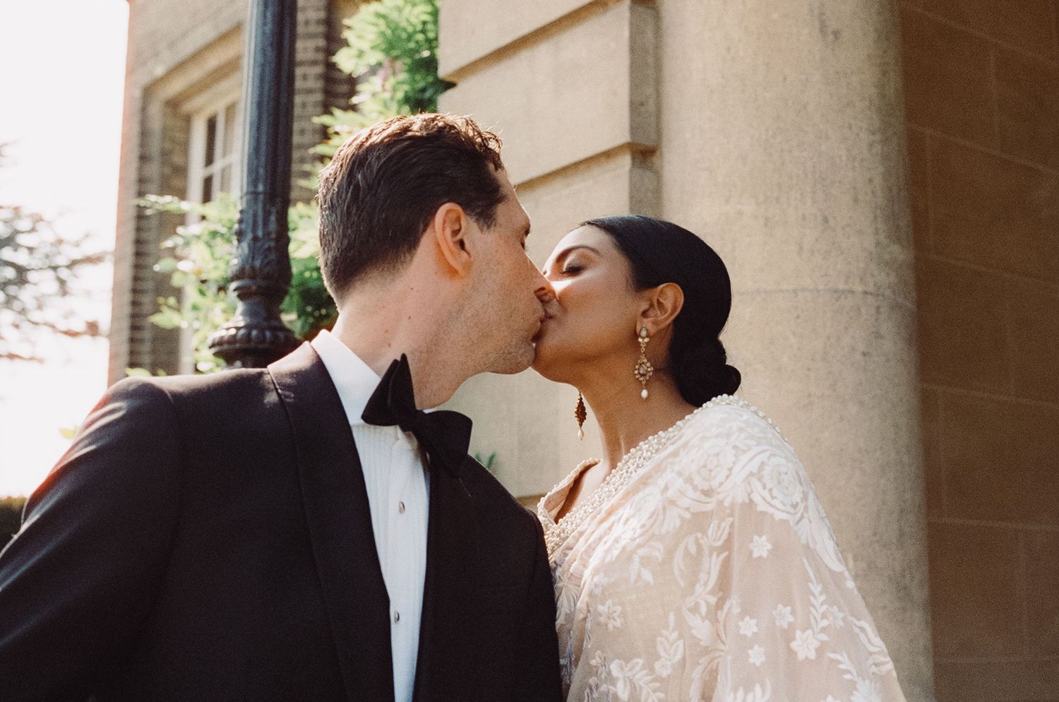 A couple shares a romantic kiss, the groom in a black tuxedo and the bride in a white lace gown, outdoors near a stone building.