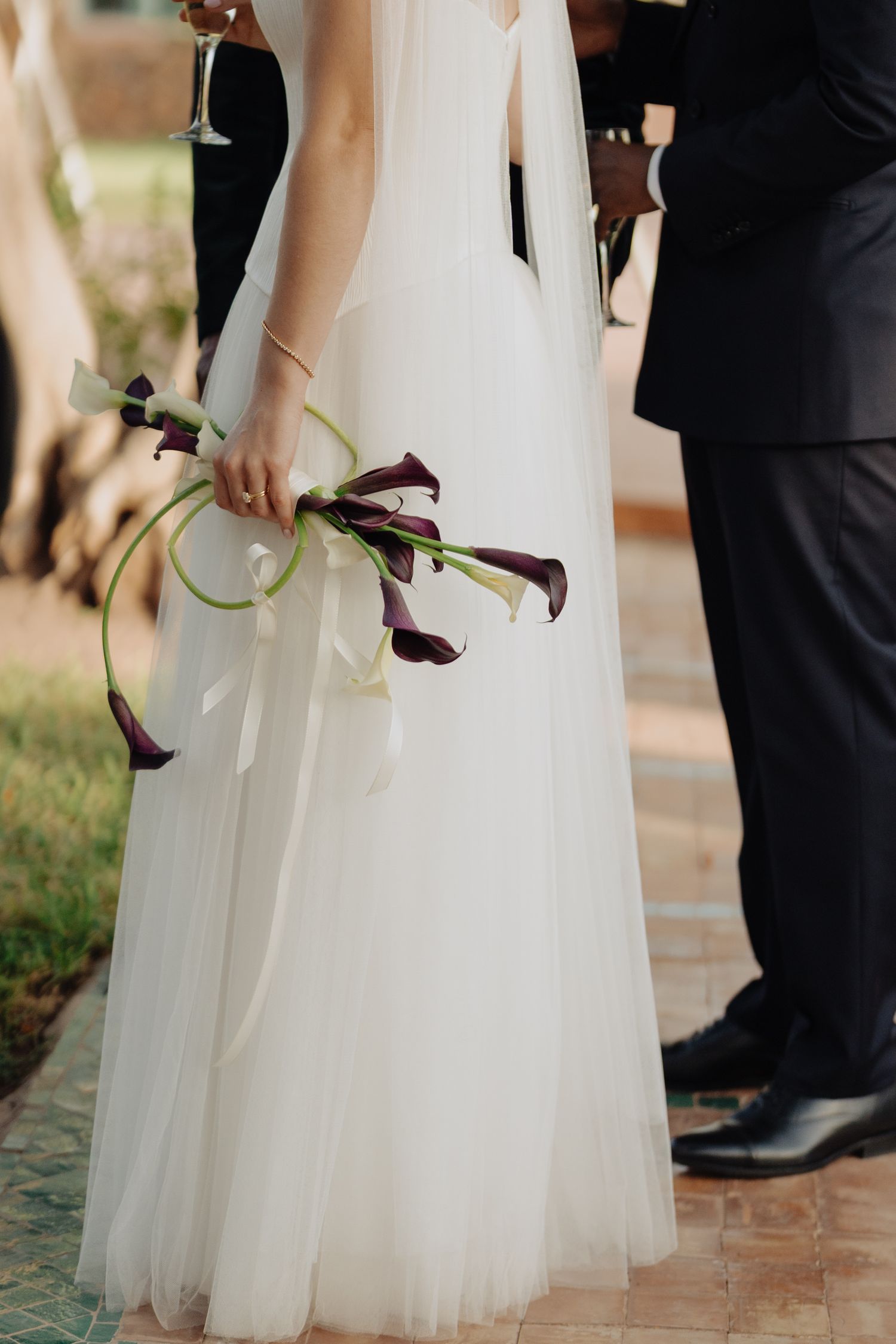 Bride in white tulle gown holding elegant dark purple calla lily bouquet at outdoor wedding ceremony.