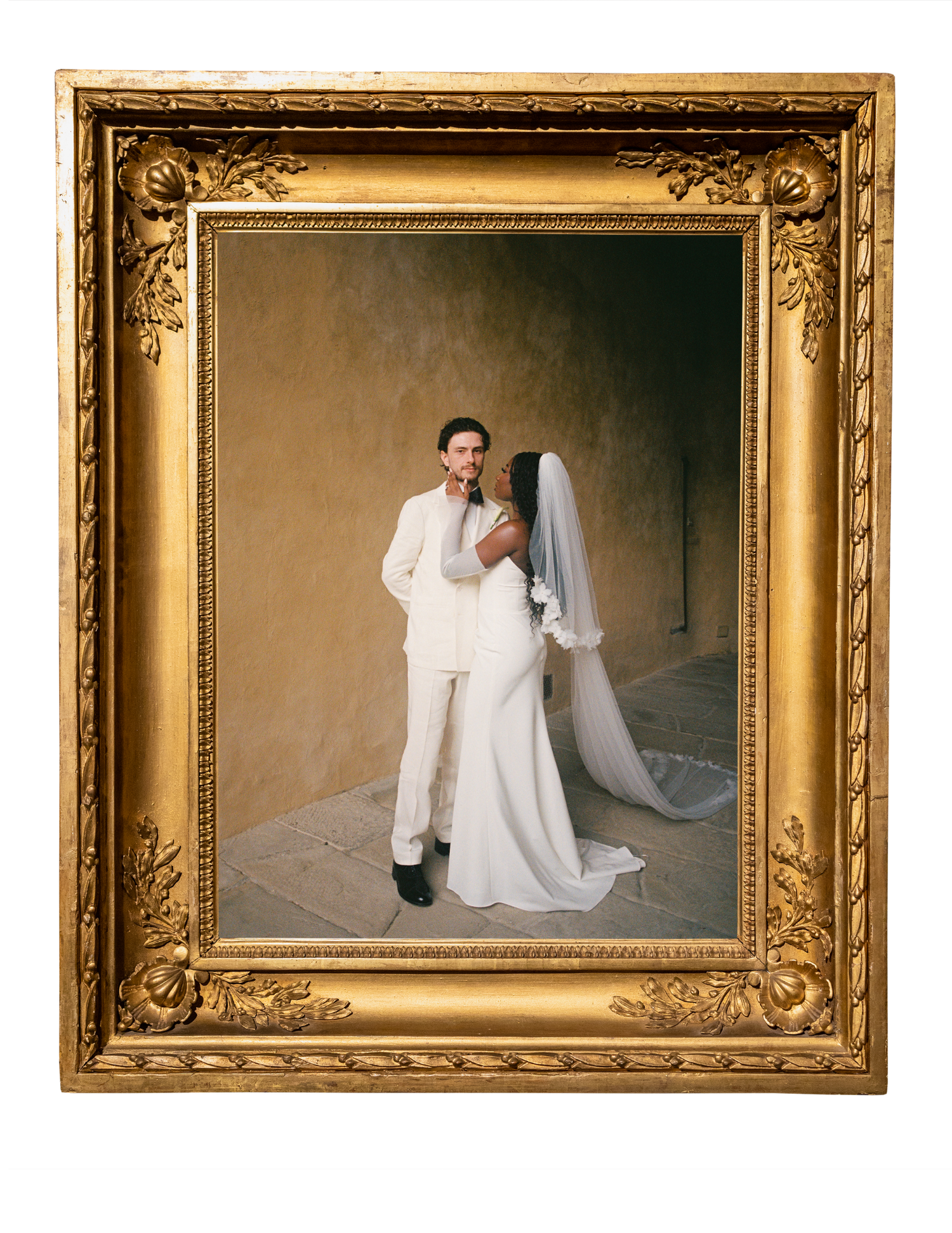 A couple in wedding attire poses in an ornate gold-framed portrait, bride in white gown with veil, groom in white suit.