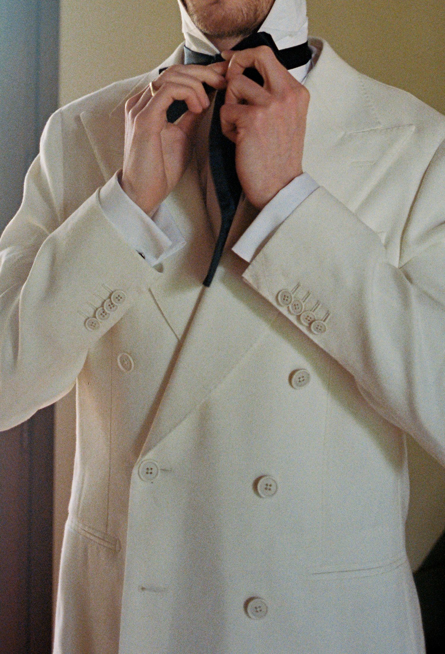 Man in white double-breasted tuxedo jacket adjusting a black bow tie, elegant formal wedding attire.