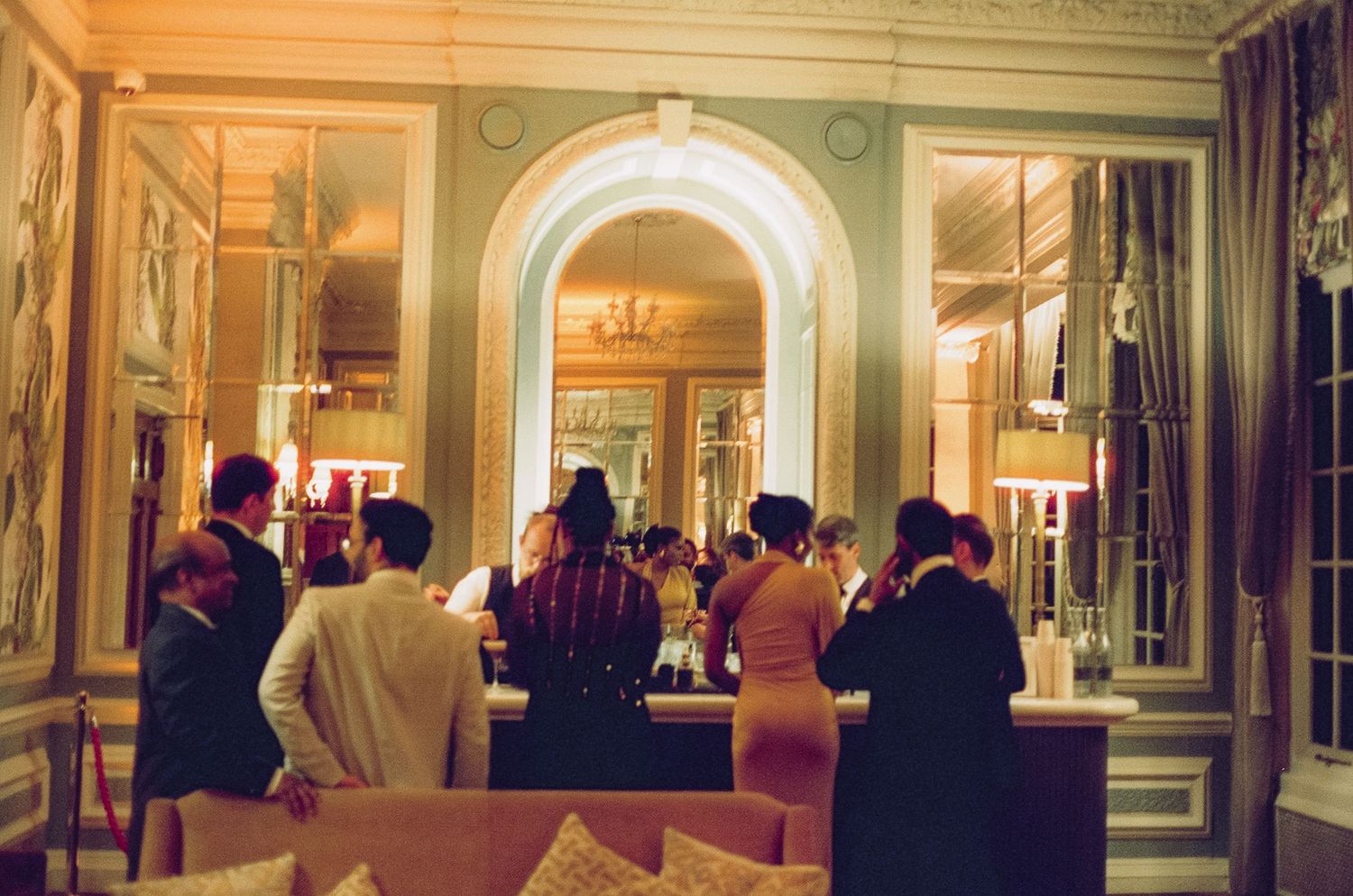 Elegant guests socializing at a luxurious bar in a grand, warmly lit classical interior with ornate archways.