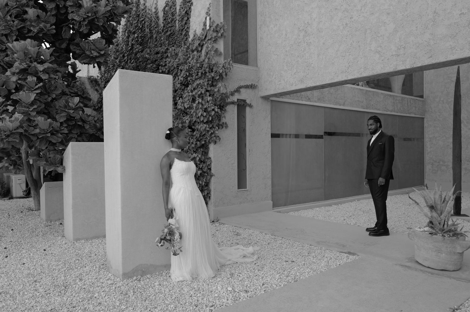 Black and white wedding photo of bride and groom standing apart in a modern courtyard with geometric pillars.