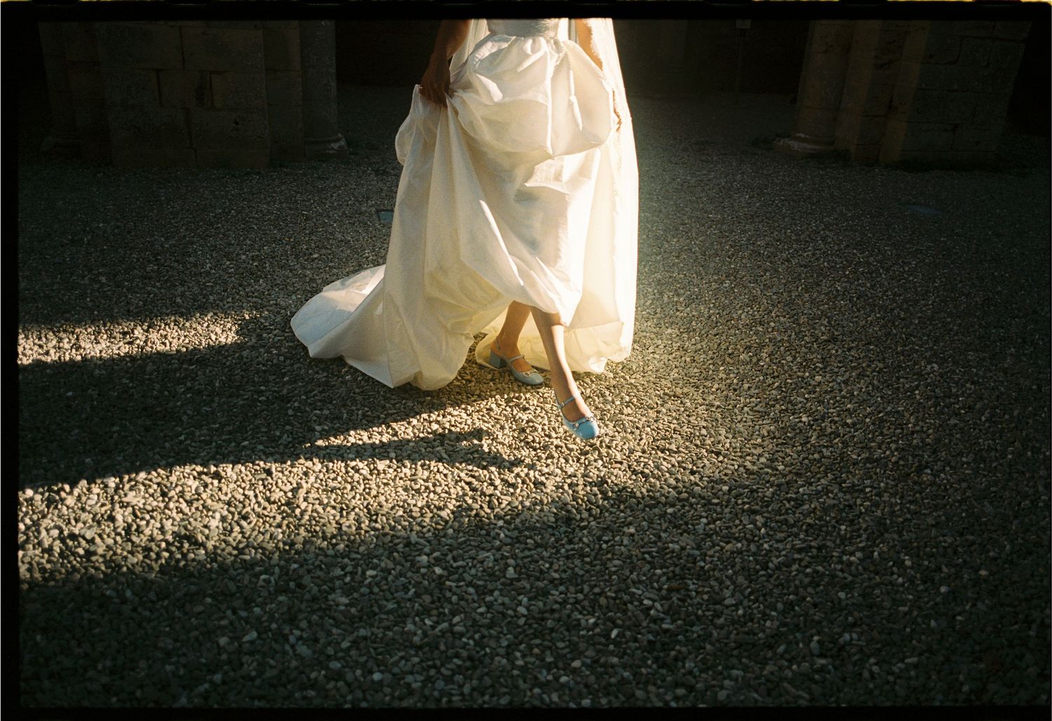 Bride in white gown walking on gravel path, sunlight casting dramatic shadows on her flowing dress and blue heels.
