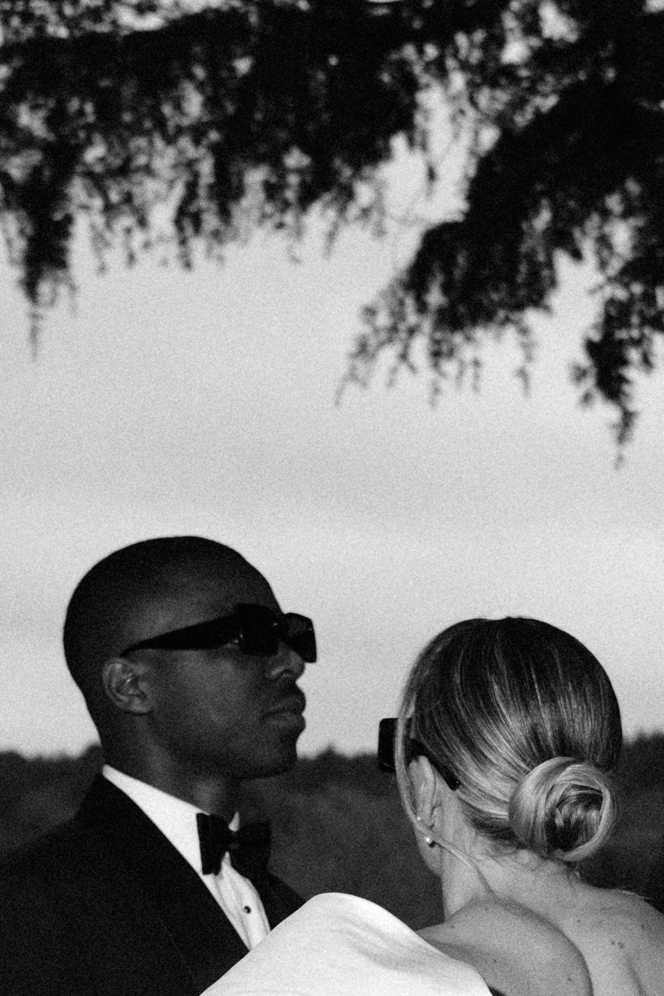 Black and white wedding photo of a couple in formal attire under a tree, groom in tuxedo and sunglasses.