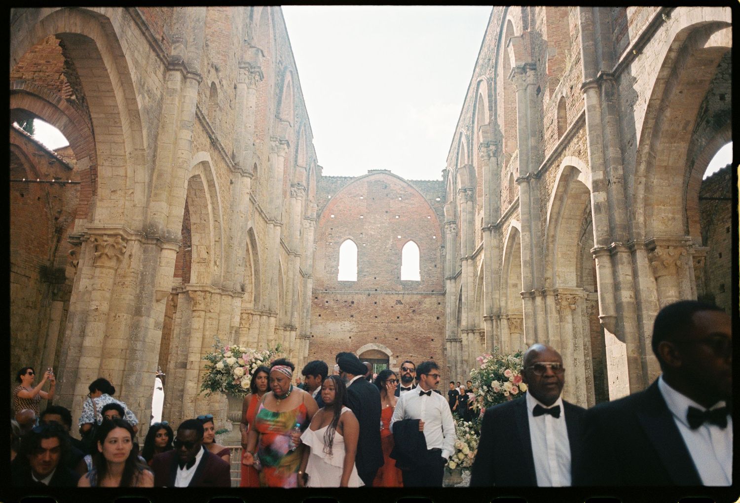 Wedding ceremony in ancient stone abbey ruins with arched columns, guests in formal attire lining the aisle.
