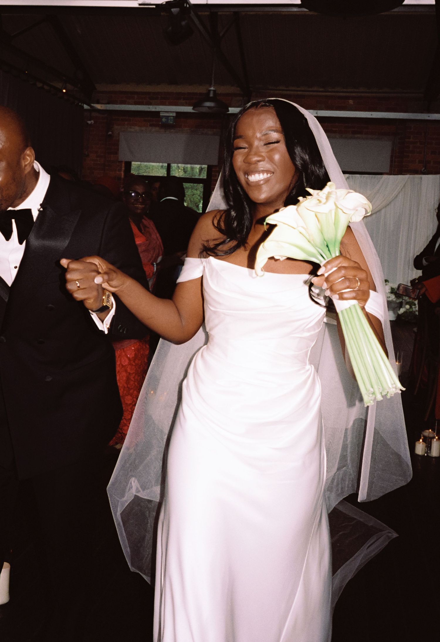 Joyful bride in white off-shoulder gown and veil holds white calla lily bouquet while dancing at wedding reception.