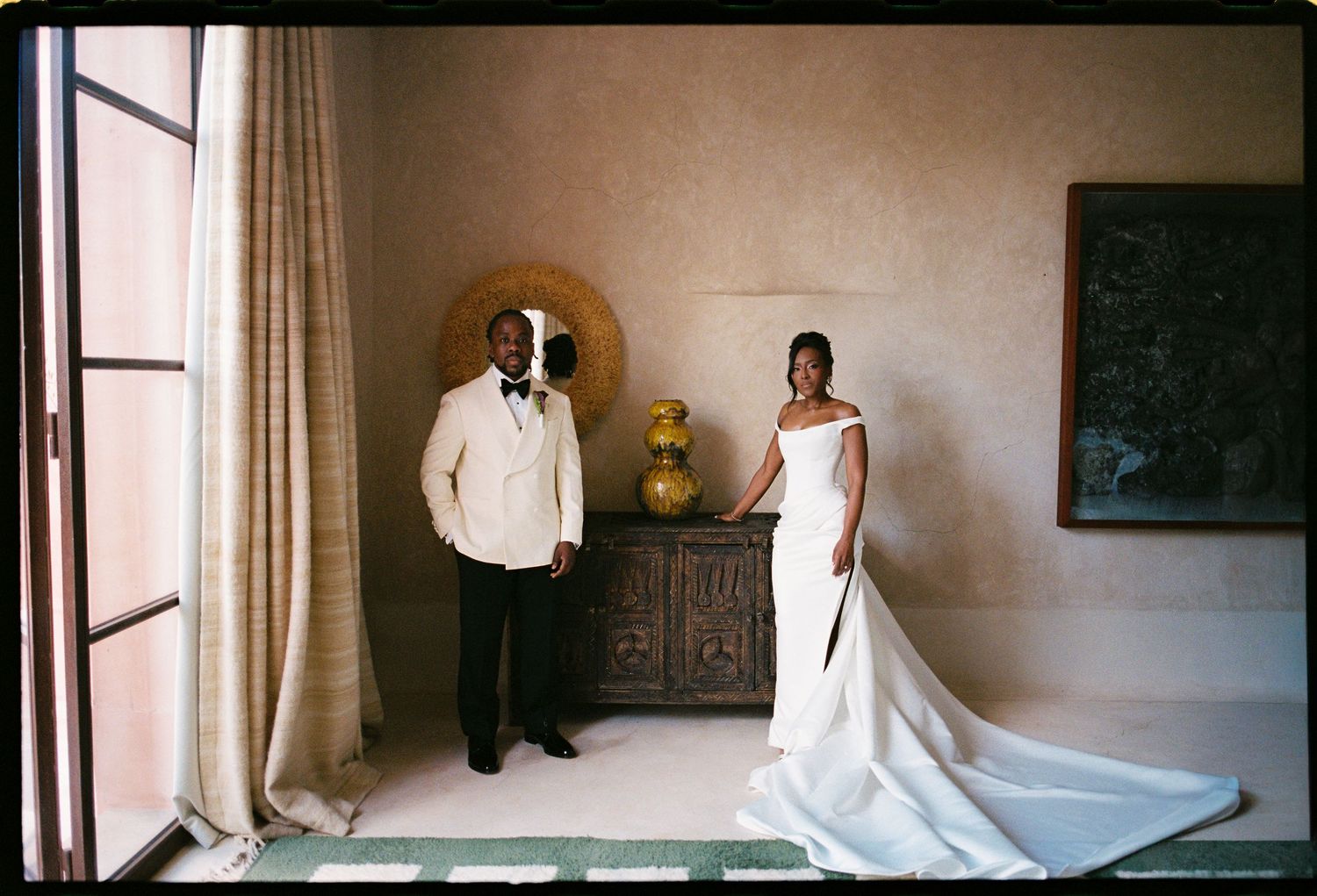 Elegant bride and groom in wedding attire posing in a warmly lit room with earthy tones and classic decor.