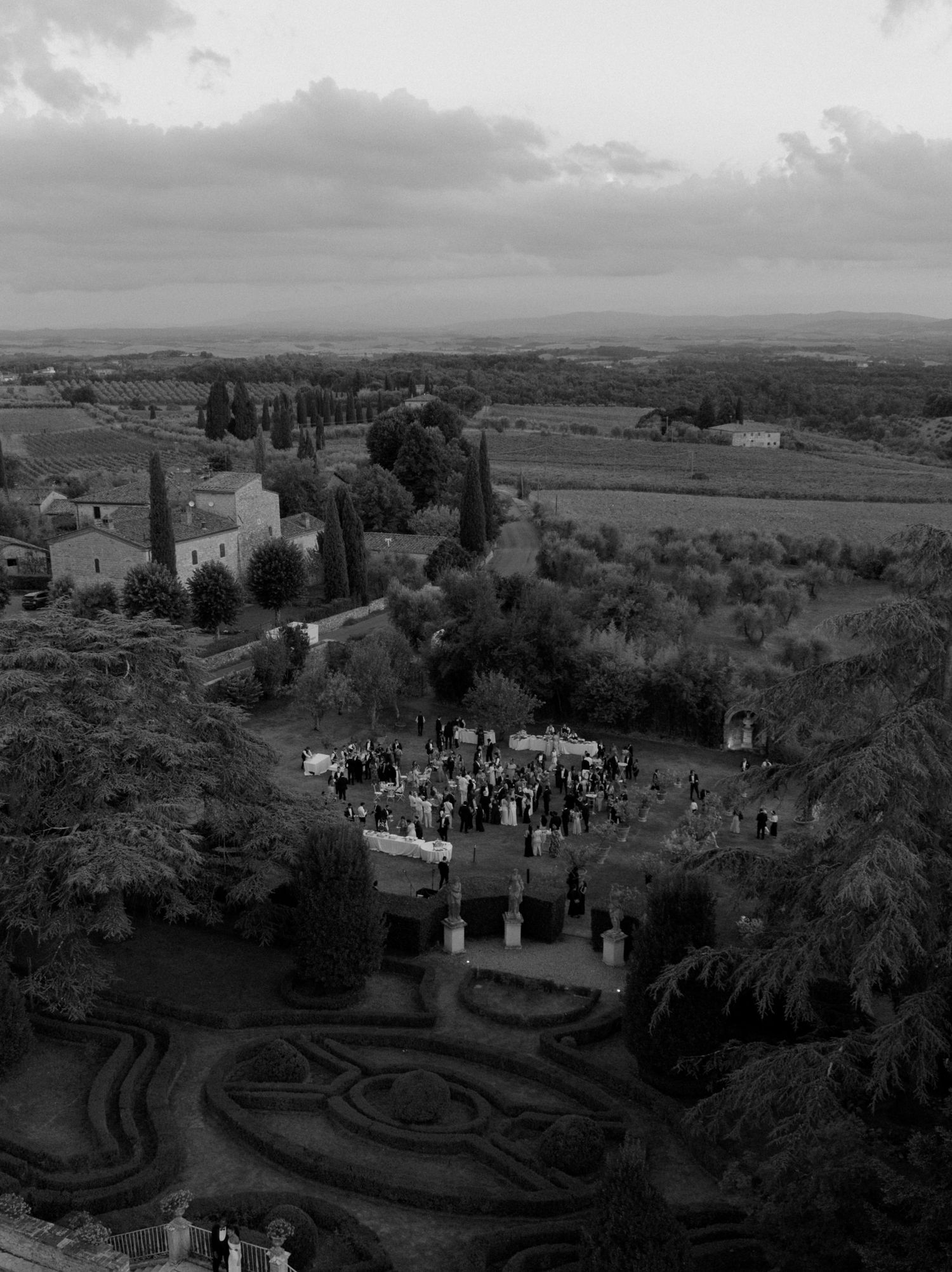 Aerial black and white view of a Tuscan estate garden with guests gathered outdoors, surrounded by cypress trees and rolling countryside.