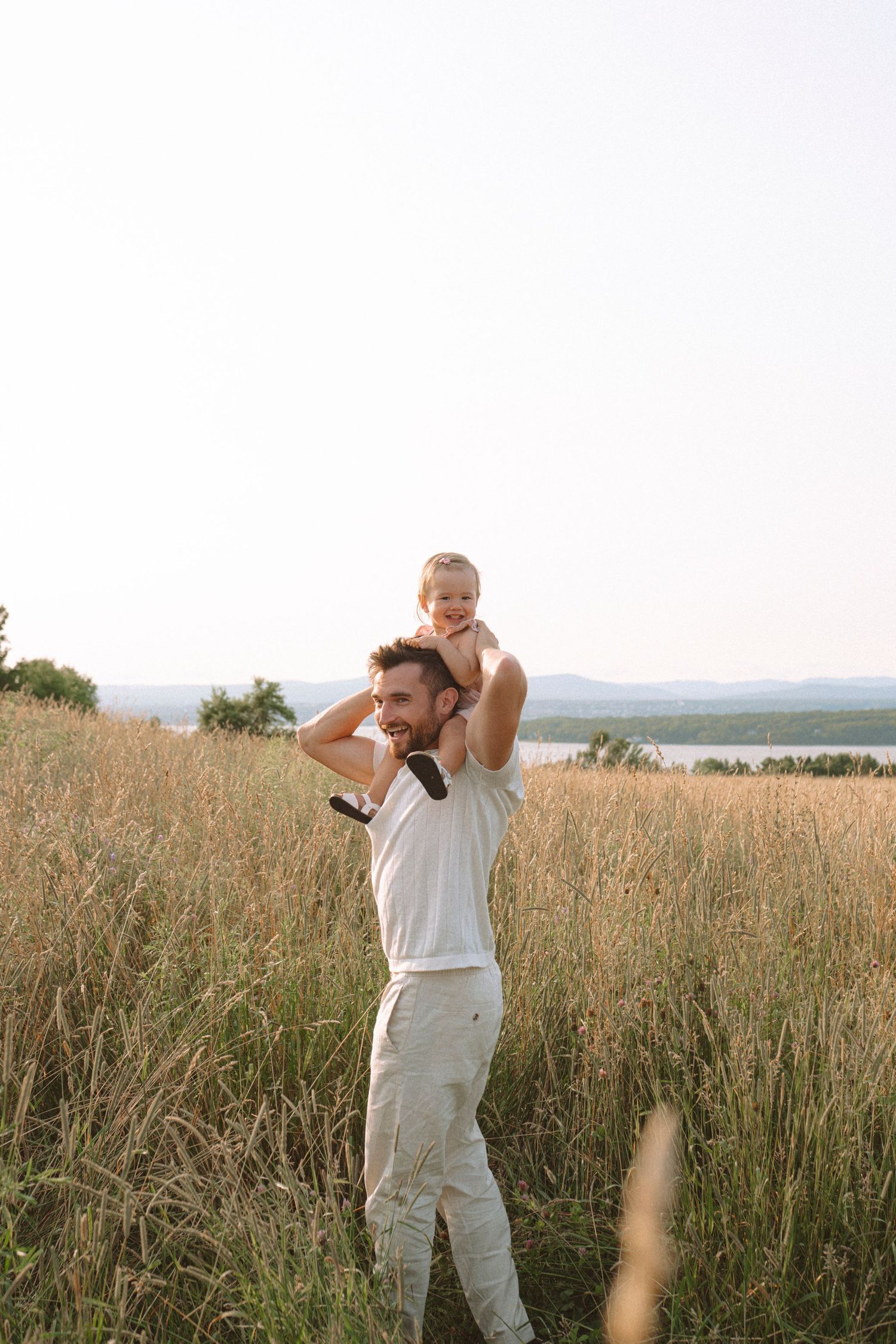 Parent and child playing together in a sun-drenched meadow overlooking water.