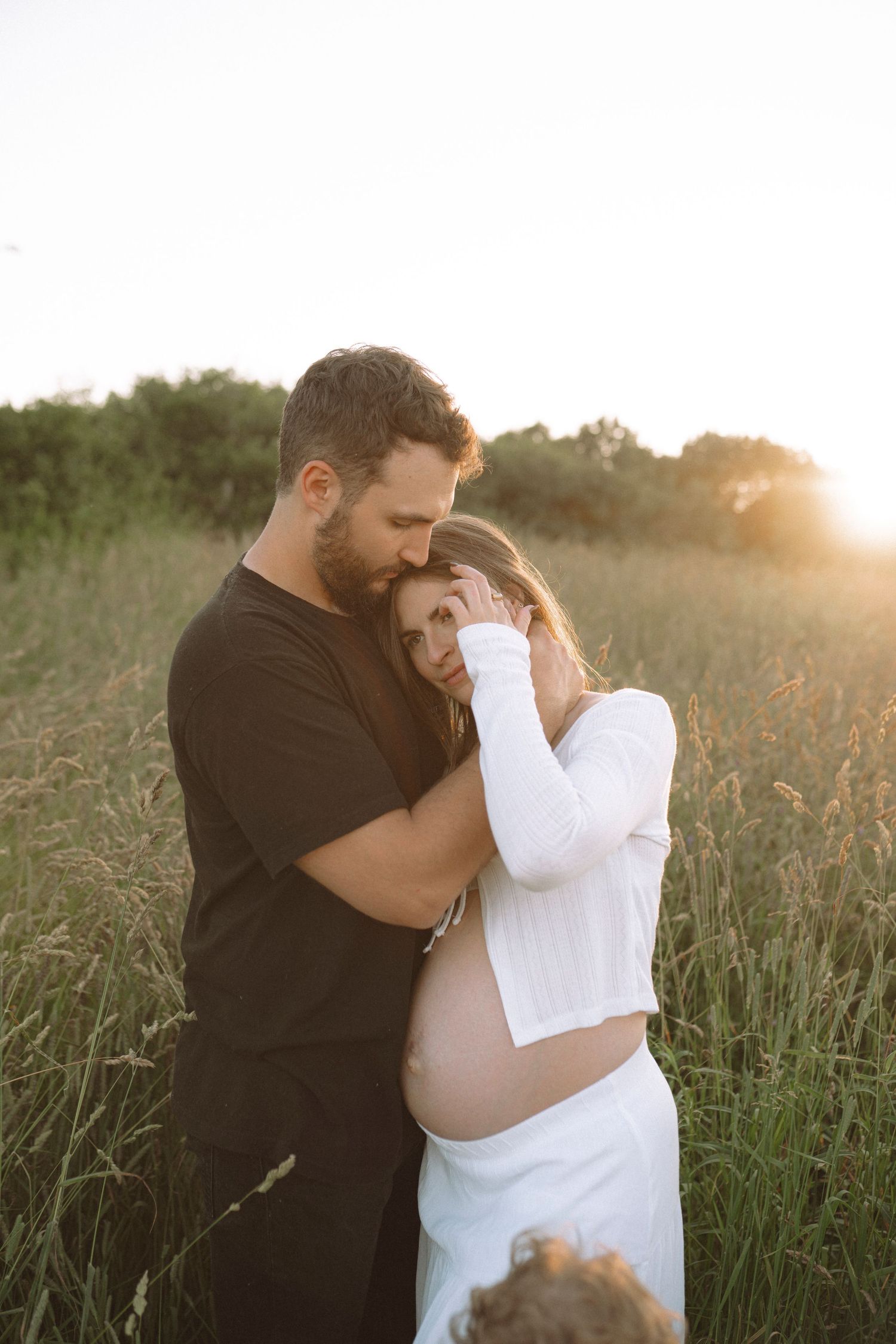 Intimate couple's moment during sunset maternity photoshoot in tall grass field.