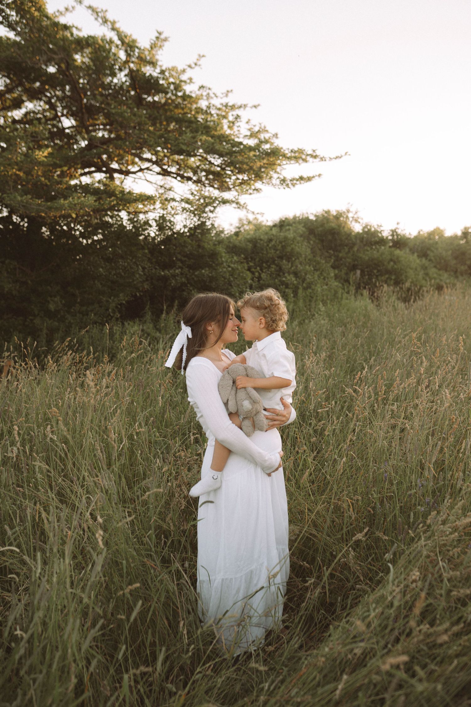Mother in flowing white dress holds toddler during golden hour photoshoot.