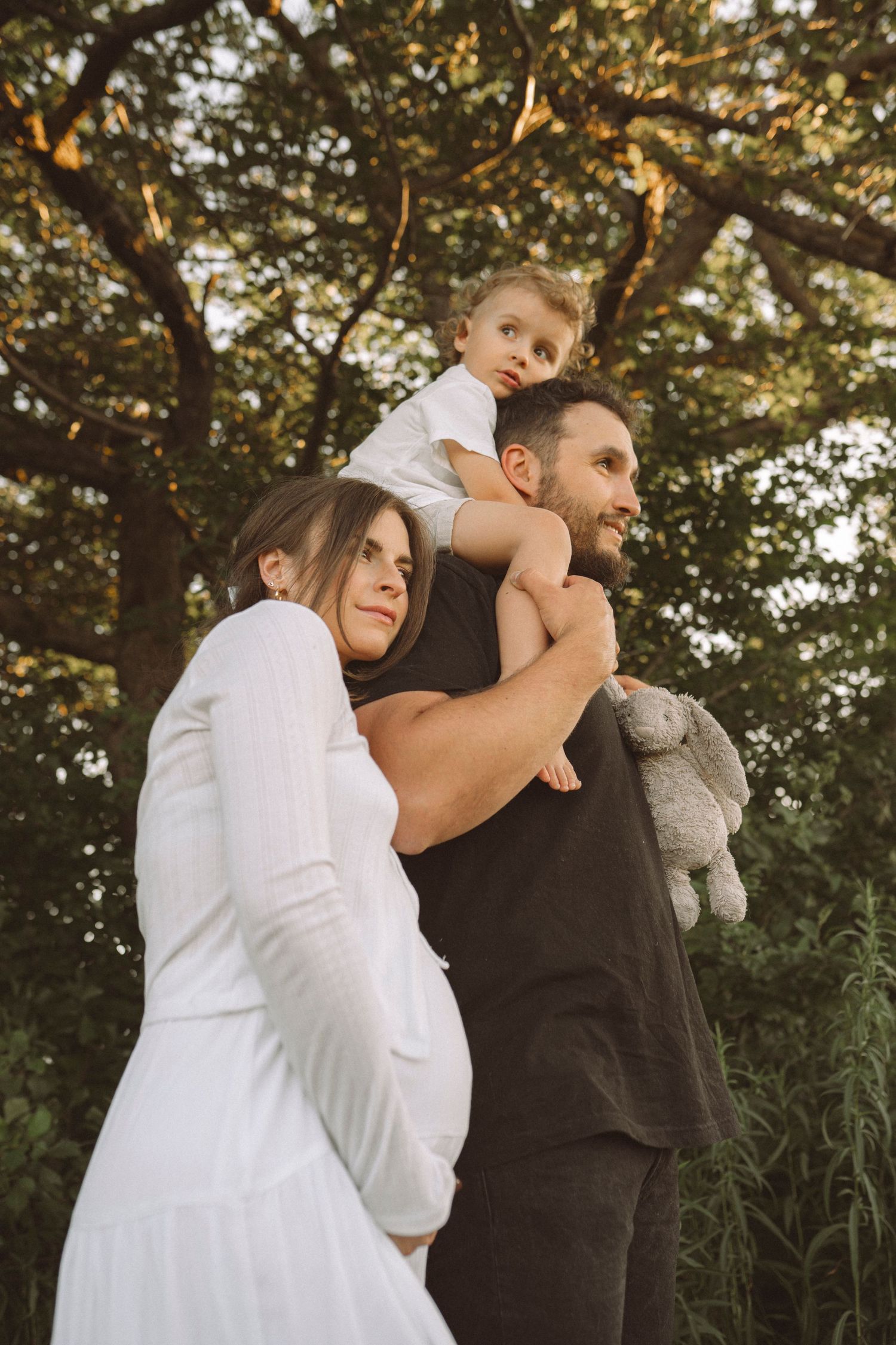 Family of three poses together in nature with child on father's shoulders.