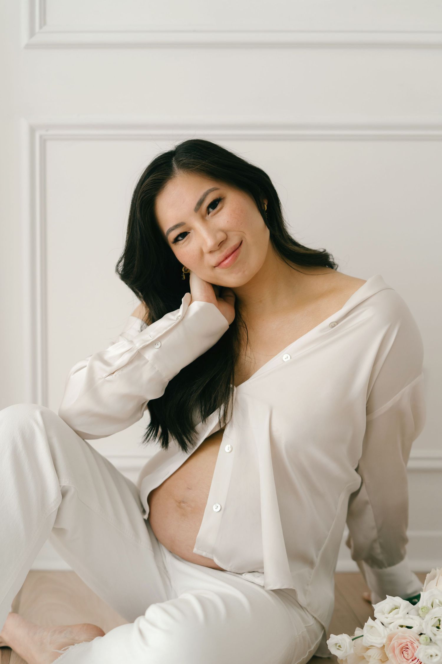 Maternity portrait of woman in white loungewear sitting against white wall.