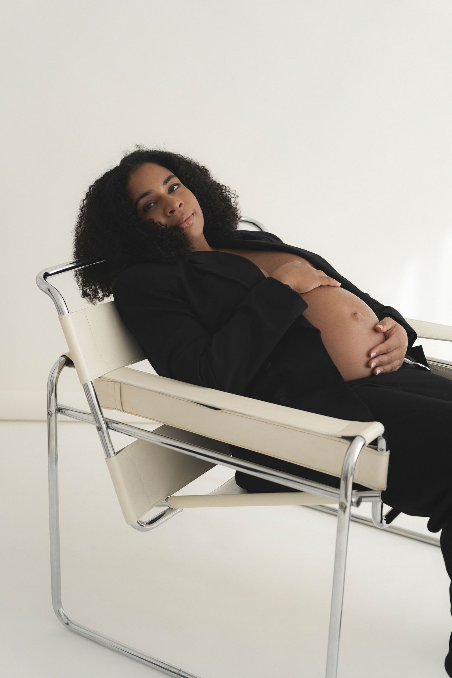 Expectant mother in black outfit reclining on modern white chair during photoshoot.