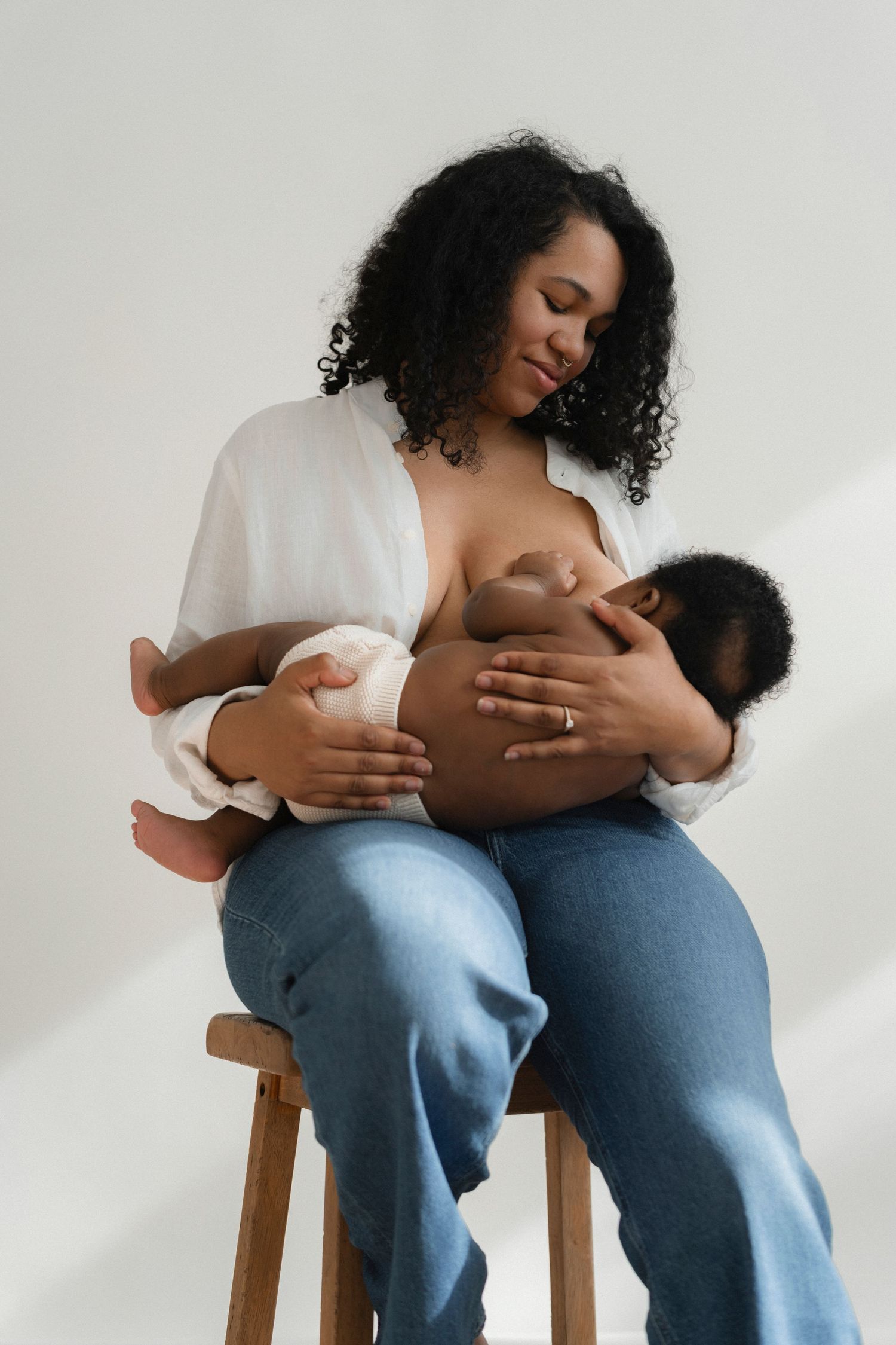 Parent in white shirt and jeans cradles nursing infant while seated on wooden stool.