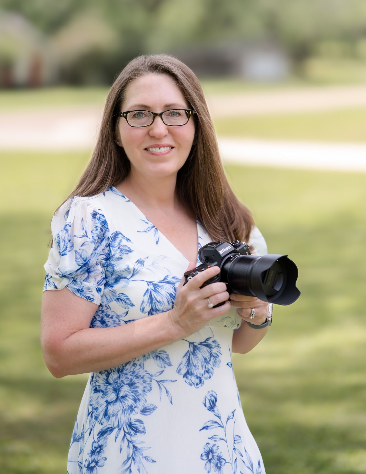 A photographer wearing a blue and white floral dress stands outdoors holding a professional camera in natural light.