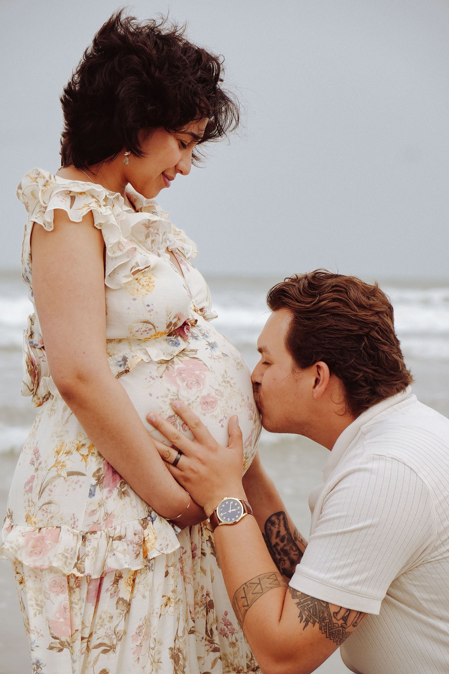 Man kneeling to kiss pregnant woman's baby bump at beach during romantic maternity photo session by ocean.