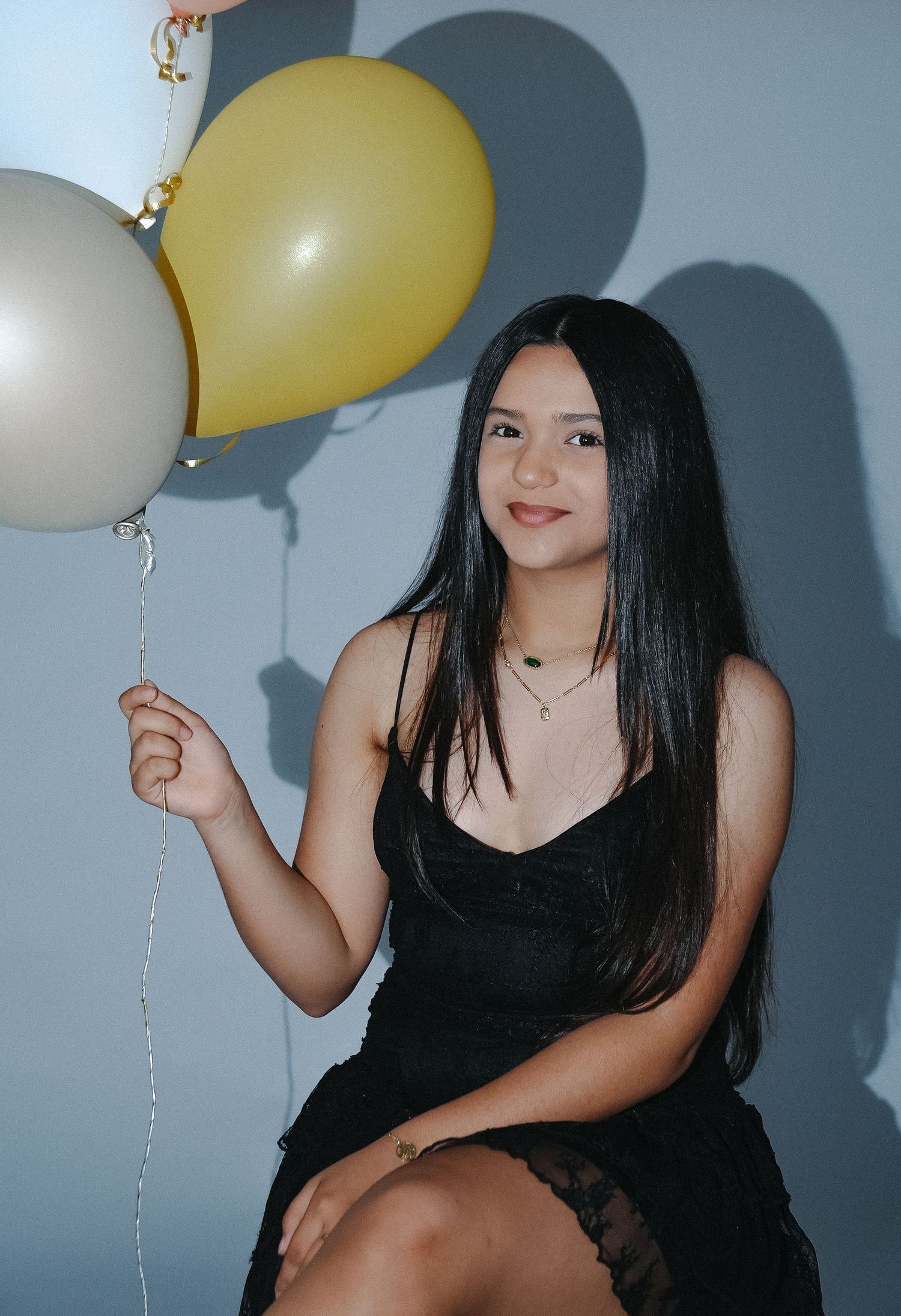Young woman in black dress holding gold and silver balloons, smiling against a gray background.