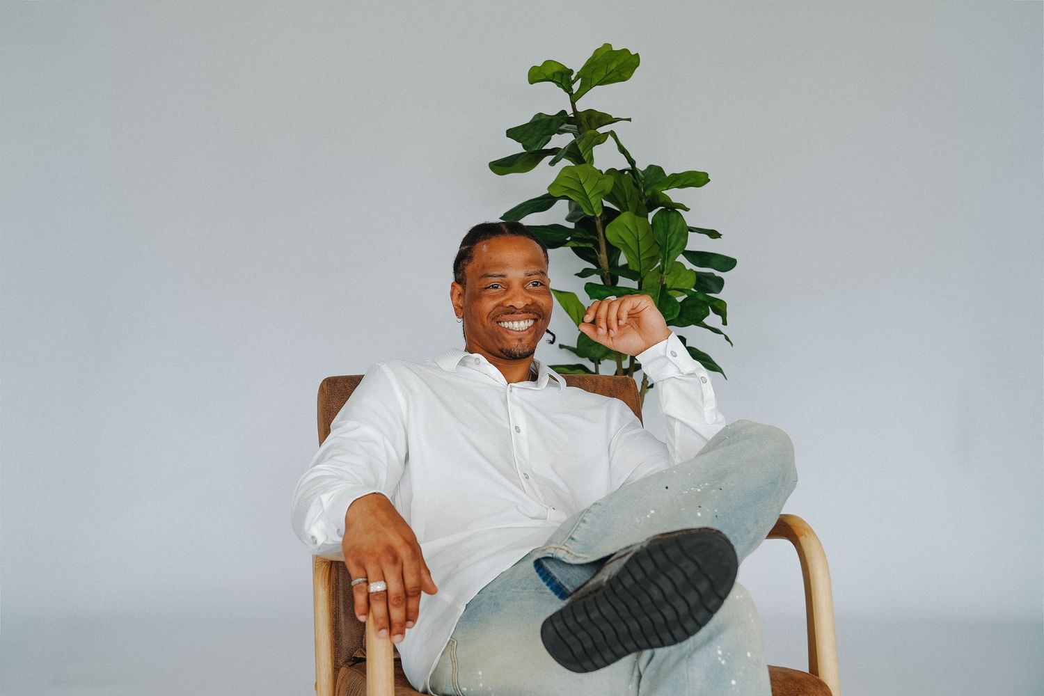 Smiling Black man in white shirt sitting cross-legged in wooden chair with green fiddle leaf fig plant behind him.