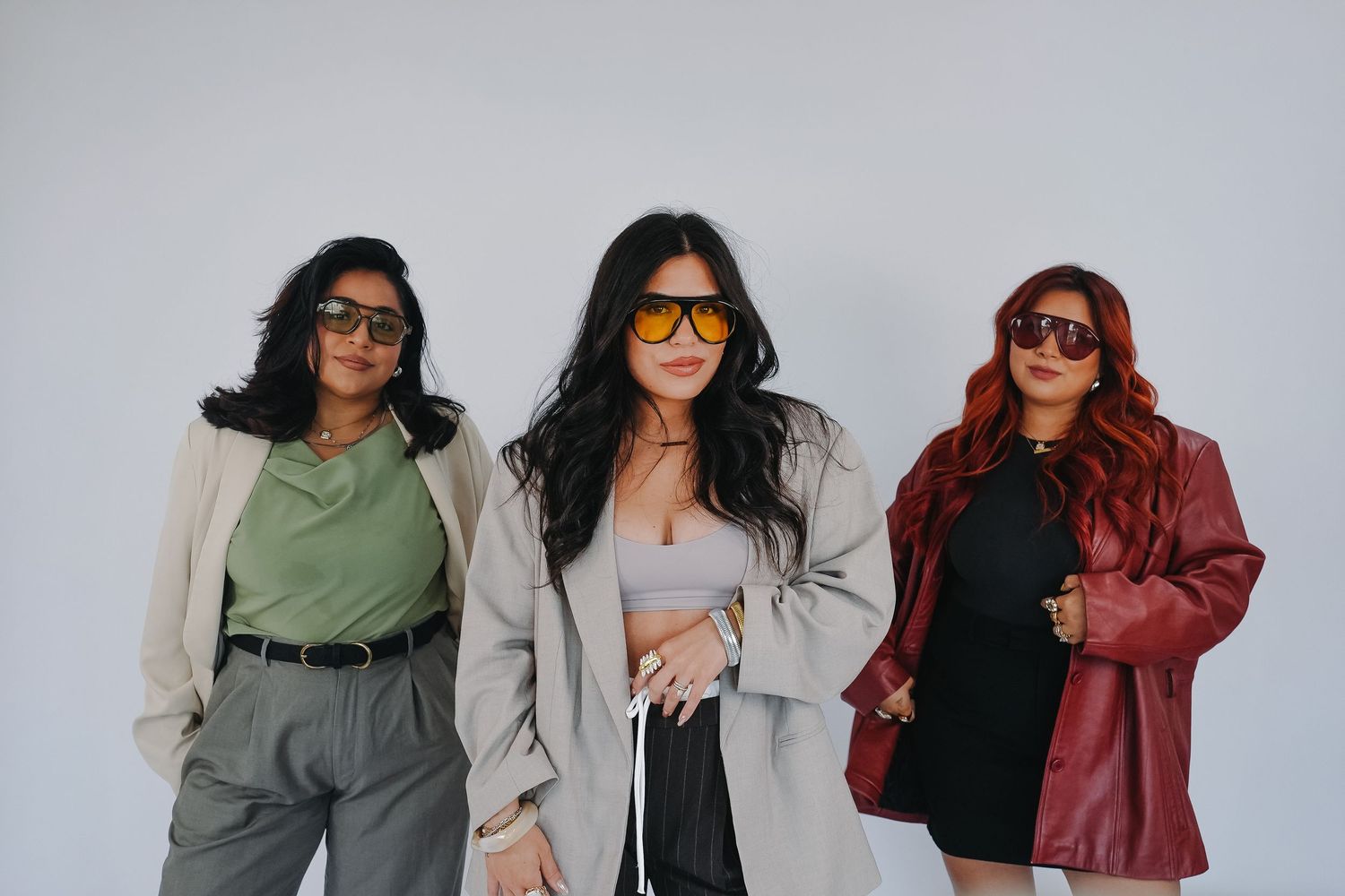 Three stylish women wearing sunglasses and trendy outfits pose confidently against a white background.